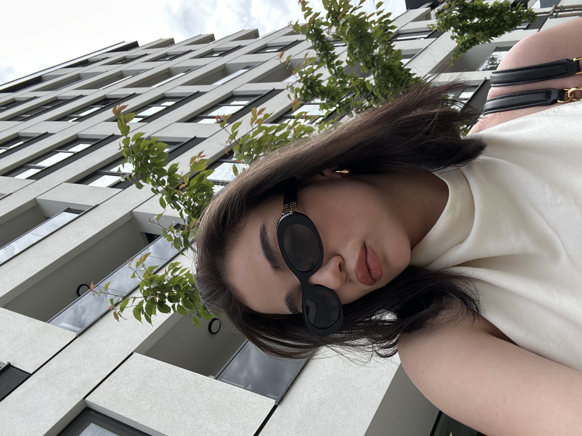 A young woman is seen from a low angle, wearing dark sunglasses and a white top, with a building facade in the background. The building has a modern design with repetitive rectangular windows. A leafy tree branch partially obscures the building and the woman. The location is likely Sofia, Bulgaria.