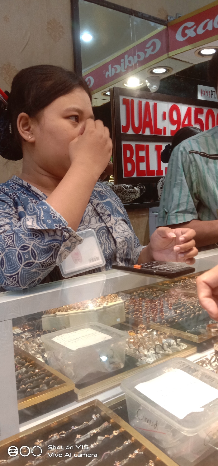 The image depicts an interior view of a gold and jewelry shop in Probolinggo, Indonesia.
**People:**
1. A woman, positioned on the left side, has dark hair pulled back and wears a blue and white batik-patterned long-sleeved shirt. Her right hand is raised, touching the area between her nose and mouth. A small, clear earring is visible in her left ear. A clear plastic ID holder or tag is clipped to the front of her shirt on the left side.
2. A second individual, partially visible on the right, wears a light green and white striped short-sleeved shirt. Their left hand is positioned on a white counter, near a black electronic calculator.
**Objects and Setting:**
- The two individuals are positioned at a white counter, which separates them from the viewpoint.
- Below the counter, multiple transparent glass display cases showcase a large quantity of rings and other small jewelry items. These items, predominantly gold-colored but also including silver and darker metals, are arranged in rectangular trays with white foam inserts and within various clear plastic containers. One container is labeled "Cimud."
- A black electronic calculator rests on the white counter between the two individuals.
- The background features a red sign with white capital letters displaying "JUAL: 9450" and "BELI," indicative of buy and sell prices for commodities like gold.
- Above this sign, a mirror reflects an inverted "Gadis" sign, also in red lettering. Faintly visible text "PT." is below the "JUAL" sign.
- The walls behind the counter are covered in light-colored wallpaper featuring a subtle repeating pattern.
- Overhead spotlights are visible in the background and reflecting in the mirror, illuminating the scene brightly.