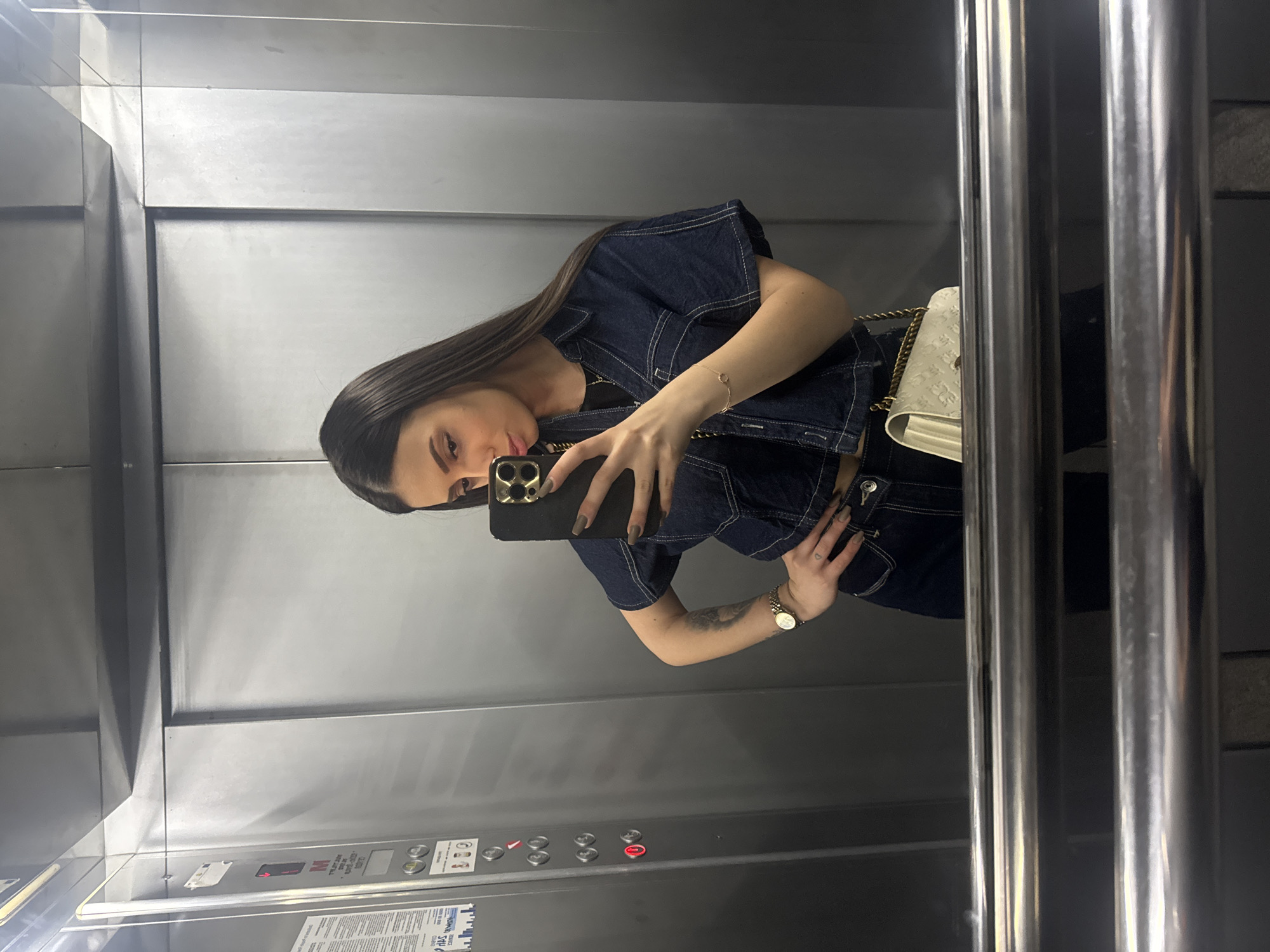 This mirror selfie, taken in an elevator in Varna, Bulgaria, features a young woman with long, straight dark hair. She is holding a black smartphone with a triple camera array to capture her reflection. The woman wears a dark denim short-sleeved top with prominent white stitching and what appears to be matching denim trousers. A light-colored, possibly quilted handbag with a gold chain strap is draped over her right shoulder.
Her left hand rests on her hip, revealing a small heart tattoo, and she wears a gold-toned watch on her left wrist. A delicate gold bracelet is visible on her right wrist. Her nails are manicured in a neutral shade. The elevator's metallic walls serve as the reflective surface for the photo. On the control panel within the elevator, various buttons are visible, including an illuminated red "M" button. Below these controls, a white informational sheet with text is present, though most of the writing is obscured by glare and angle, with only fragmented letters such as "HAS" vaguely discernible. The lighting is consistent with an indoor, artificially lit environment.