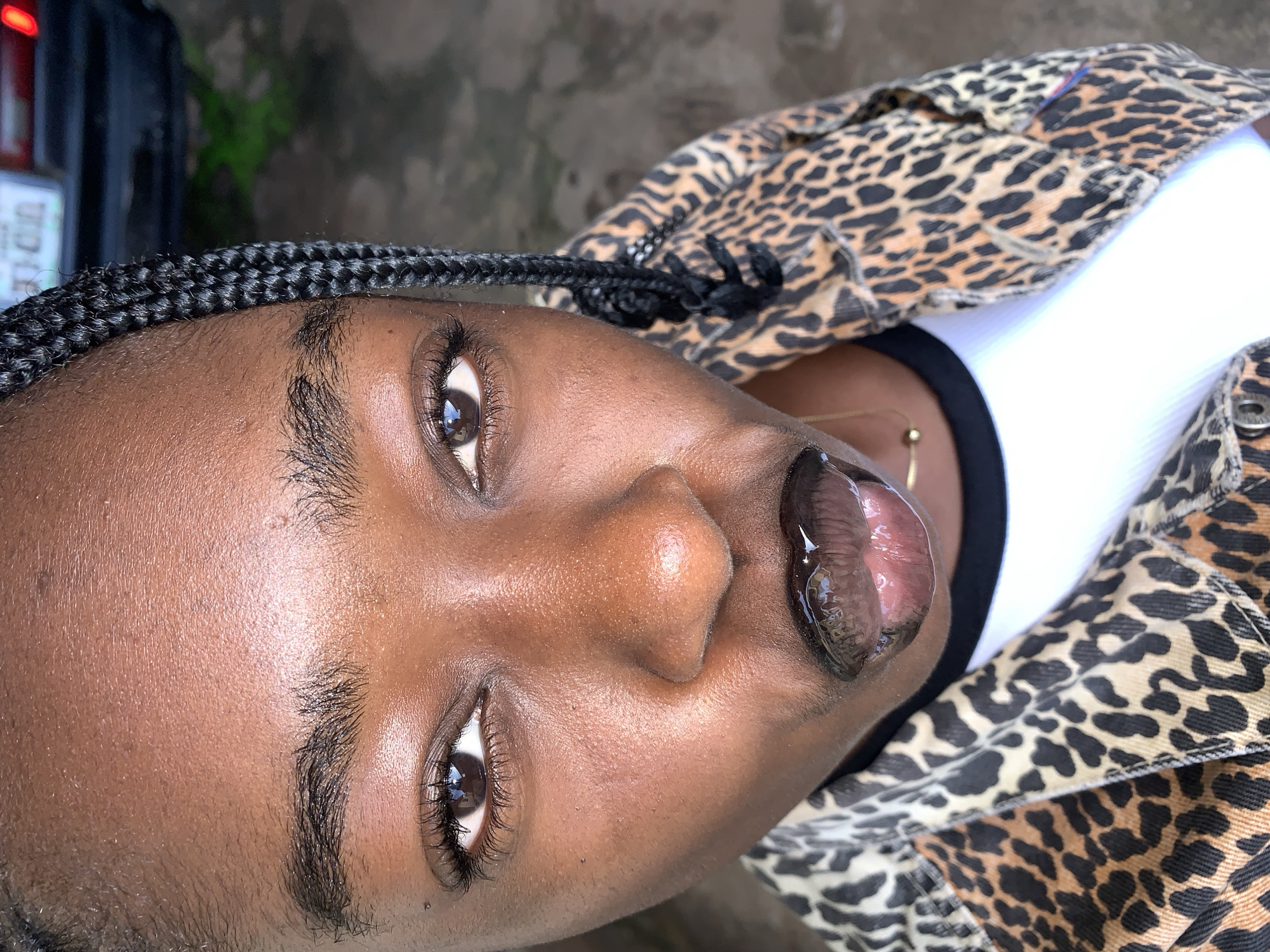A close-up, overhead shot captures a young Black woman with dark skin, sporting a leopard-print jacket over a white t-shirt, and wearing dark lipstick. Her hair is styled in braids, with one braid framing her face. She is looking directly at the camera with her mouth slightly open, revealing a glossy finish on her lips. A thin gold necklace with a single bead is visible around her neck. The background is blurred, showing an outdoor environment with what appears to be a road and some greenery. The scene is likely in the city of Rafin Par, Nigeria.