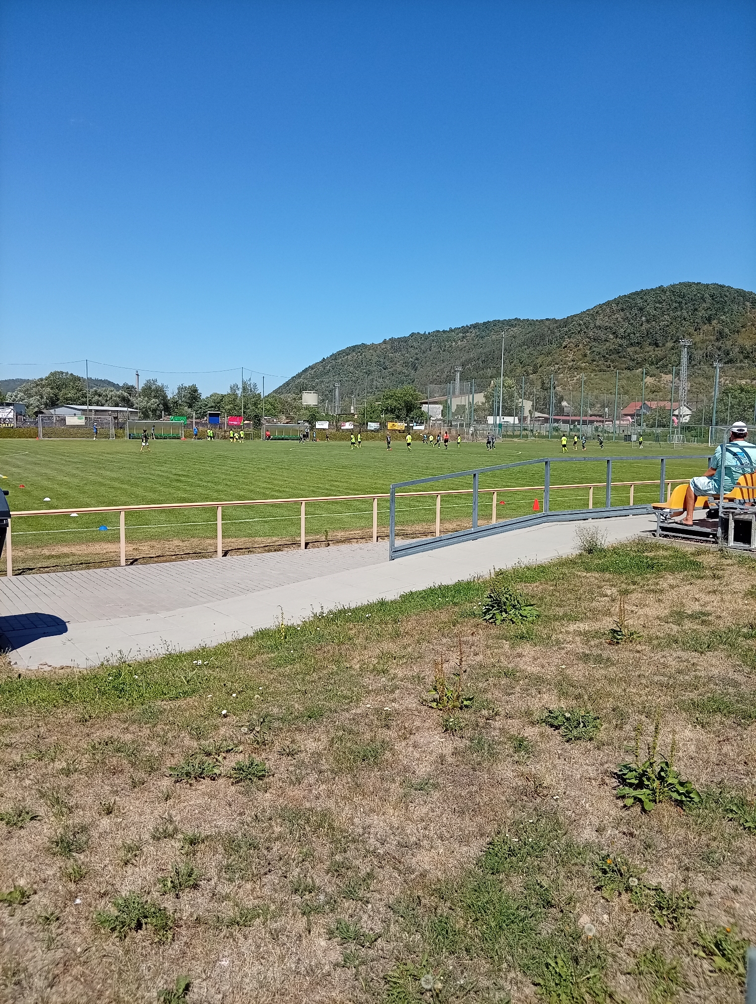 The image shows a view of a football field in the city of Tišnov, Czechia, on a clear, sunny day. The field is green and well-maintained, with several players in yellow and black uniforms visible practicing. There are goalposts at either end of the field. In the foreground, there is a dry, grassy area and a paved walkway with a metal railing. A person is sitting on a bench to the right, facing the field. In the background, there are trees and buildings, with a hilly, forested landscape beyond. The sky is a clear, bright blue.