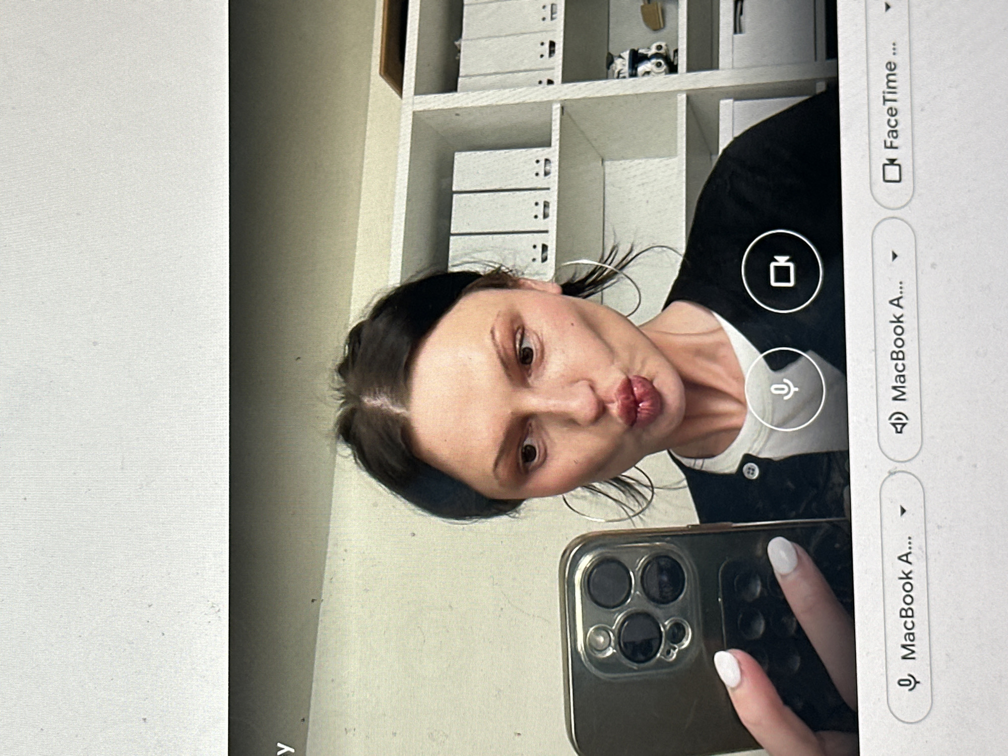 A screen displays a partial view of a woman engaged in a video call, simultaneously taking a selfie with a mobile phone. The woman, with dark hair parted in the middle and wearing large hoop earrings, has her lips puckered. She is dressed in a white t-shirt under a black cardigan. Her left hand, with white polished nails, holds a silver/gold smartphone featuring a multi-lens camera system, reflecting light. The background behind her consists of a plain light-colored wall on the left and a white shelving unit on the right containing several white binders, some adorned with simple smiley face drawings. The screen interface shows microphone and camera icons, along with text indicating 'MacBook A...' and 'FaceTime...'. The depicted scene is located in the city of Wealden, United Kingdom.