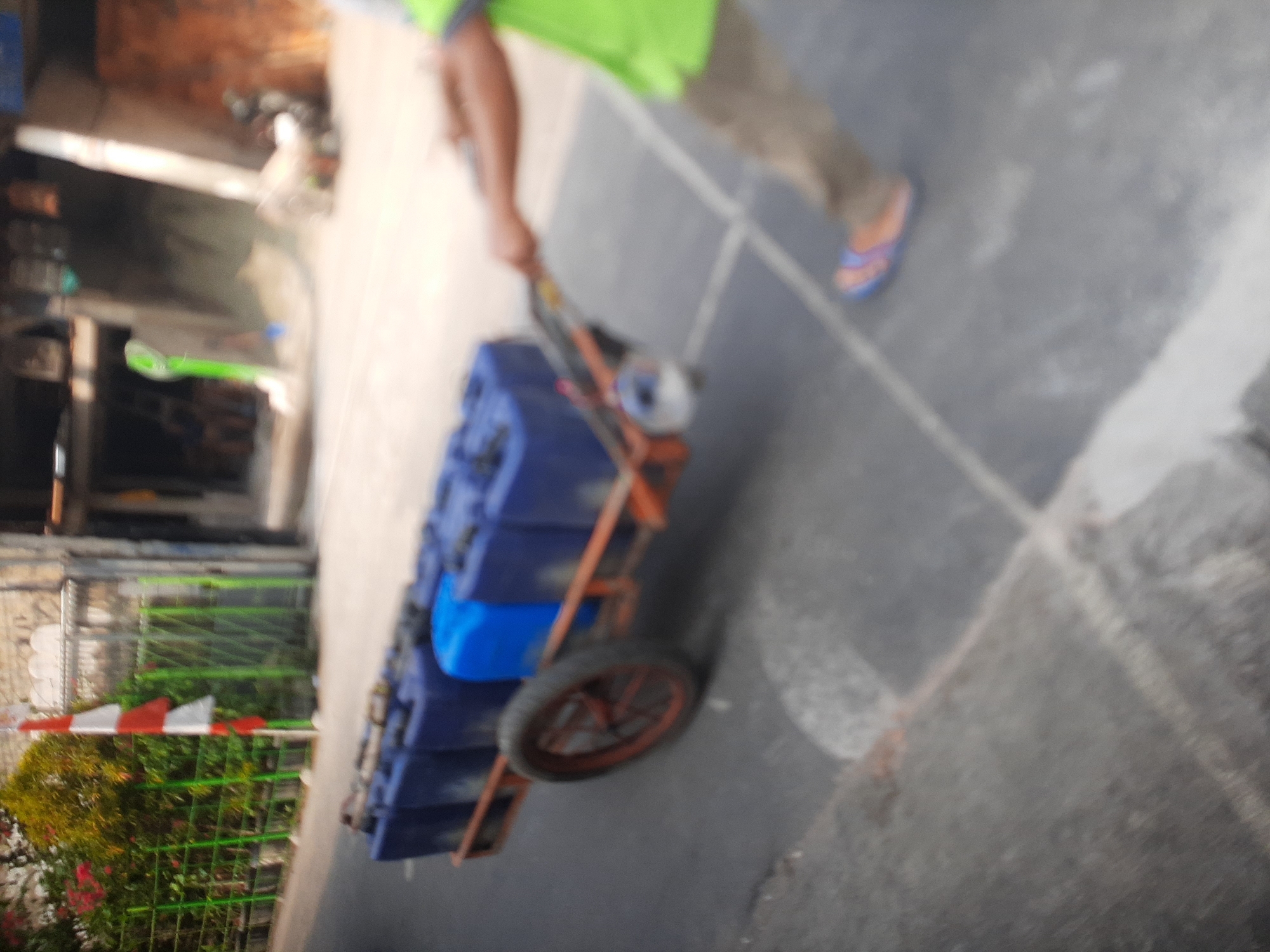 In Central Jakarta, Indonesia, a person is pulling a cart laden with blue plastic containers along a paved road. The cart has an orange metal frame and a single wheel. The person is wearing a green vest and brown pants, with blue and orange sandals. In the background, there is a white and red Indonesian flag and some greenery behind a fence. Buildings are visible further in the background. The scene appears to be outdoors on a street.