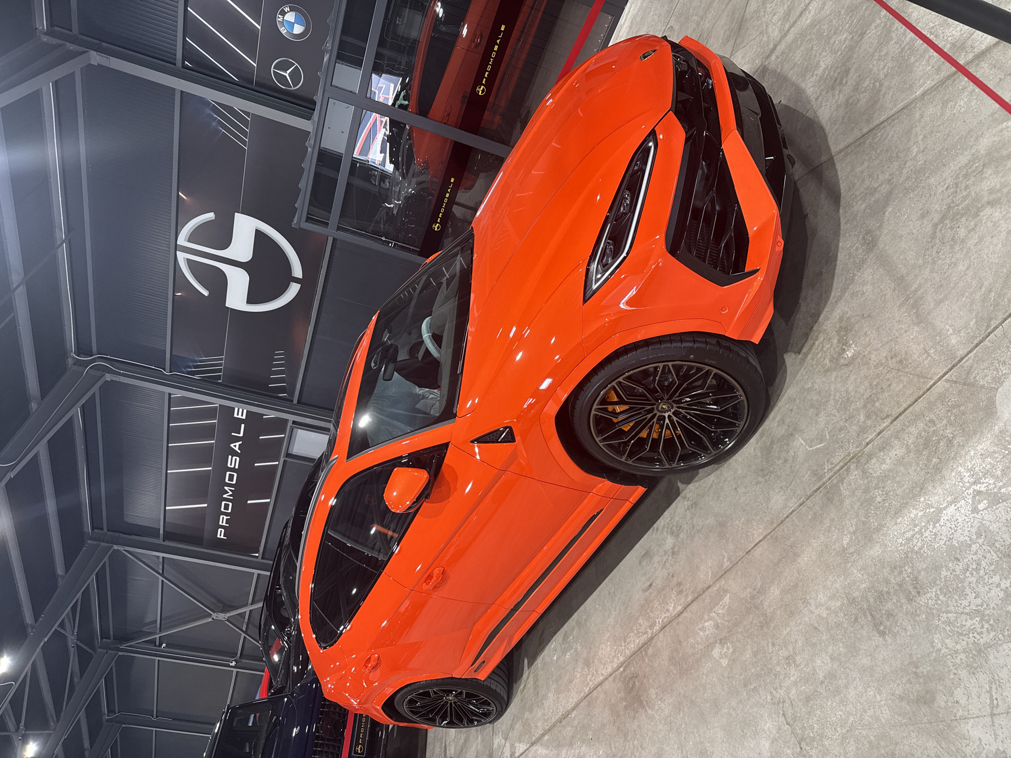 The image shows an orange Lamborghini Urus parked on a grey concrete floor in what appears to be a car showroom or dealership. The showroom has a black wall with the logos of BMW, Mercedes-Benz, and another company (shaped like a letter "D" split with a "H"). The word "PROMOSALE" is written vertically on the wall. The vehicle is angled towards the right of the image, and a portion of another vehicle, dark blue or black, is visible in the lower left corner. The environment appears to be well-lit, and the setting is indoors. The photo was taken in Plovdiv, Bulgaria.