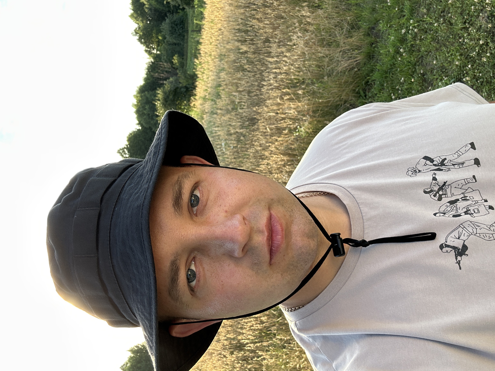 A medium shot captures a man from the chest up, wearing a black bucket hat and a light-colored t-shirt. The t-shirt has a graphic design of cartoon-like figures holding weapons. A black drawstring with a toggle closure hangs from the hat under the man's chin. The man's head is slightly tilted and he is looking directly at the camera. His eyes are open, and his expression is neutral.
The background consists of a field of dry grass illuminated by the setting sun, creating a golden hue. Beyond the field, a line of green trees forms a treeline against a bright, possibly hazy, sky. The lighting suggests it is late afternoon or early evening. The location is identified as Dubyshche, Ukraine.