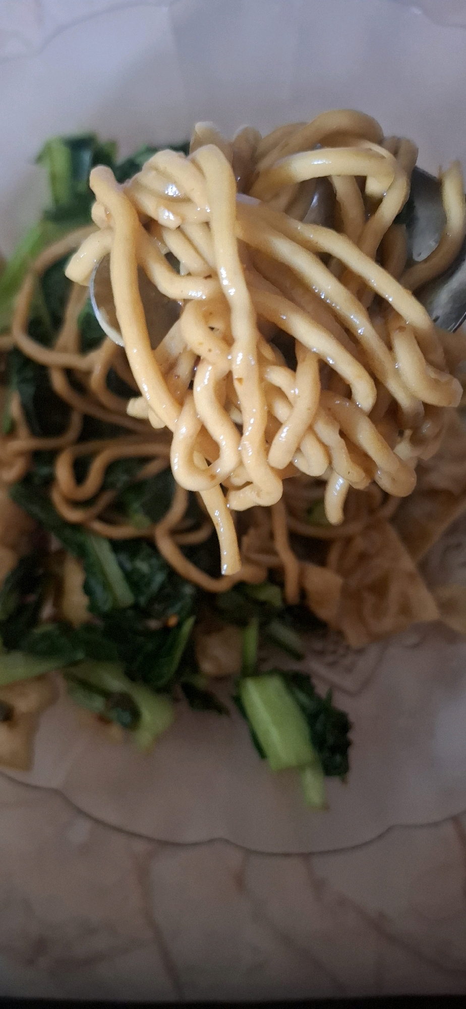 In Ciharashas, Indonesia, a close-up shot focuses on a fork lifting a portion of stir-fried noodles. The noodles, a pale yellow, are coated in a light sauce and intermingled with chopped green vegetables, likely bok choy or similar leafy greens. A few pieces of fried wonton or similar crispy fried dough are visible underneath the noodles. The background is blurred, but suggests a casual dining setting, possibly a home or a small eatery. The lighting appears to be natural or ambient, casting soft shadows.