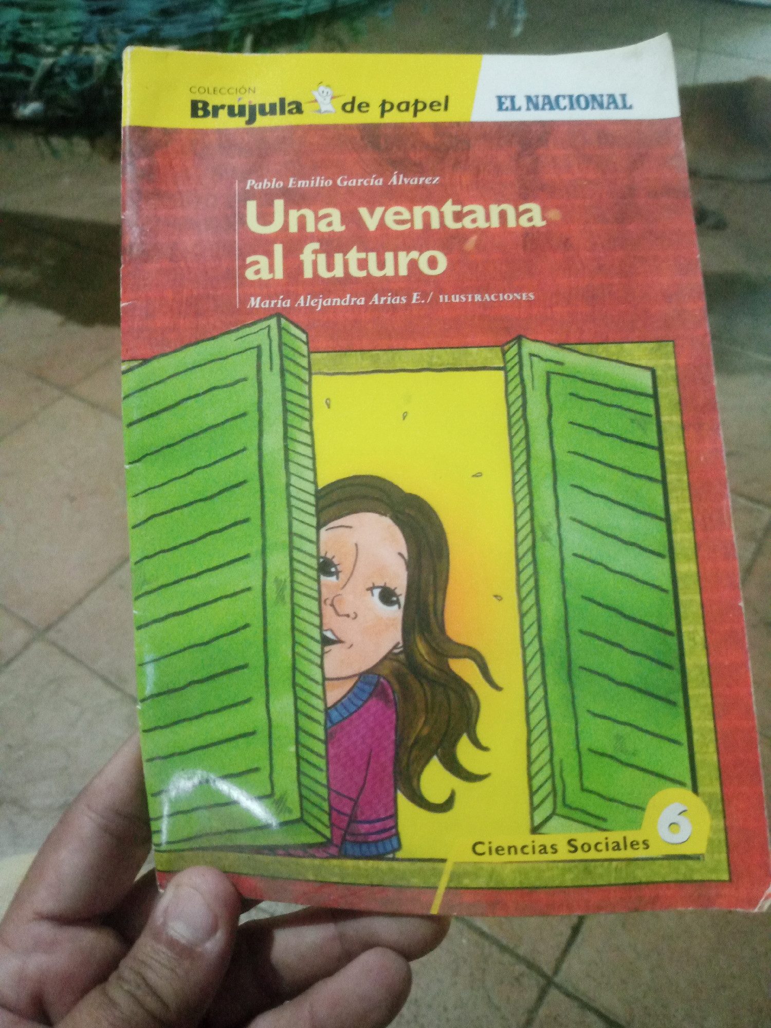 A book cover is held in a person's left hand. The book title reads "Una ventana al futuro" (A window to the future). The cover features a drawing of a girl peeking out from behind a green shuttered window. The background is yellow and red. The book appears to be part of a collection called "Brujula de papel" and published by "El Nacional". The bottom right corner of the cover indicates "Ciencias Sociales" (Social Sciences) and the number "6". The image was taken in Santa Ana, Venezuela.