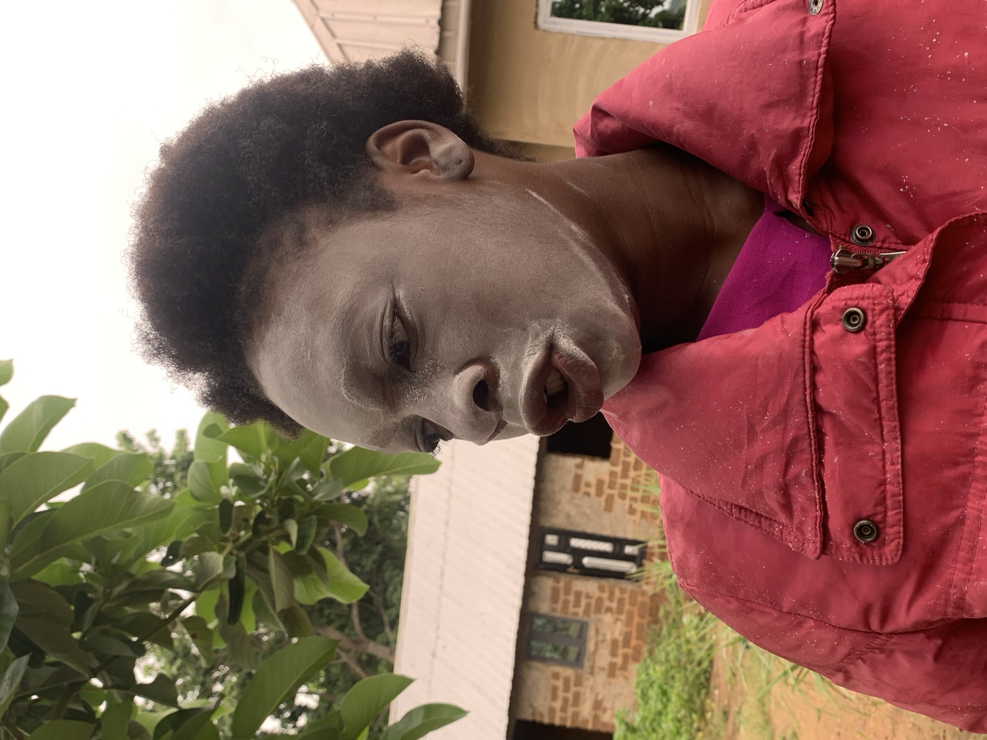 A medium shot, taken outside in Agban, Nigeria, depicts a person with dark skin covered in a pale, possibly powdery, substance on their face and neck. They are wearing a red jacket over a purple garment. The individual is positioned near a lush green tree with large leaves, and behind them is a building with light-colored walls and dark window frames. The building appears to be constructed of brick in some areas. The scene is overcast.