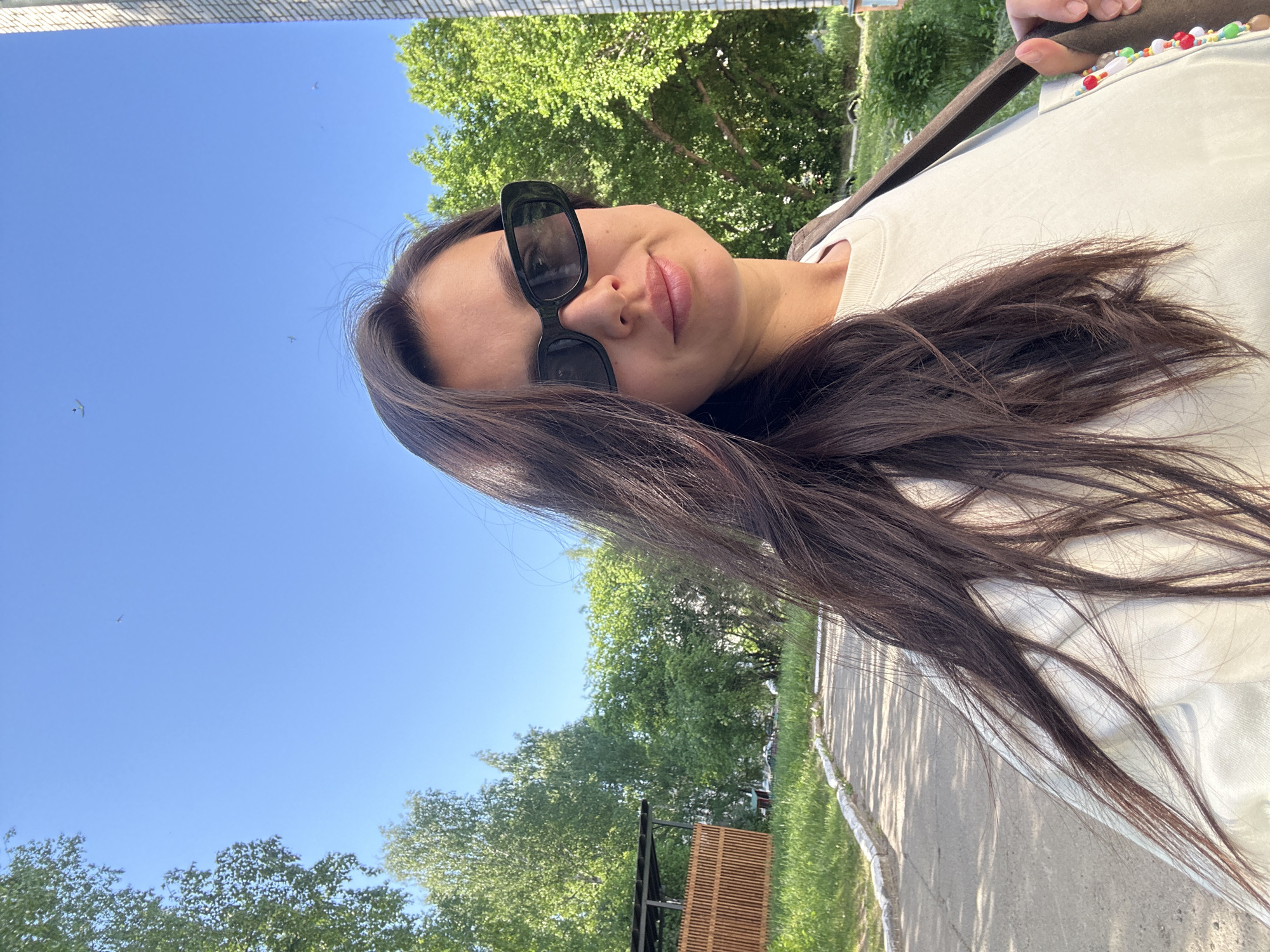 A woman with long, dark hair and sunglasses is facing the camera. She is wearing a light-colored shirt with a beaded strap or decoration visible on the shoulder. The background consists of a clear blue sky and lush green trees. A paved pathway and some sort of wooden structure can be seen in the distance. The image suggests an outdoor setting in a park-like area. There is not enough information in the image to determine if the photo was taken in the city of Chaikovsky, Russia.