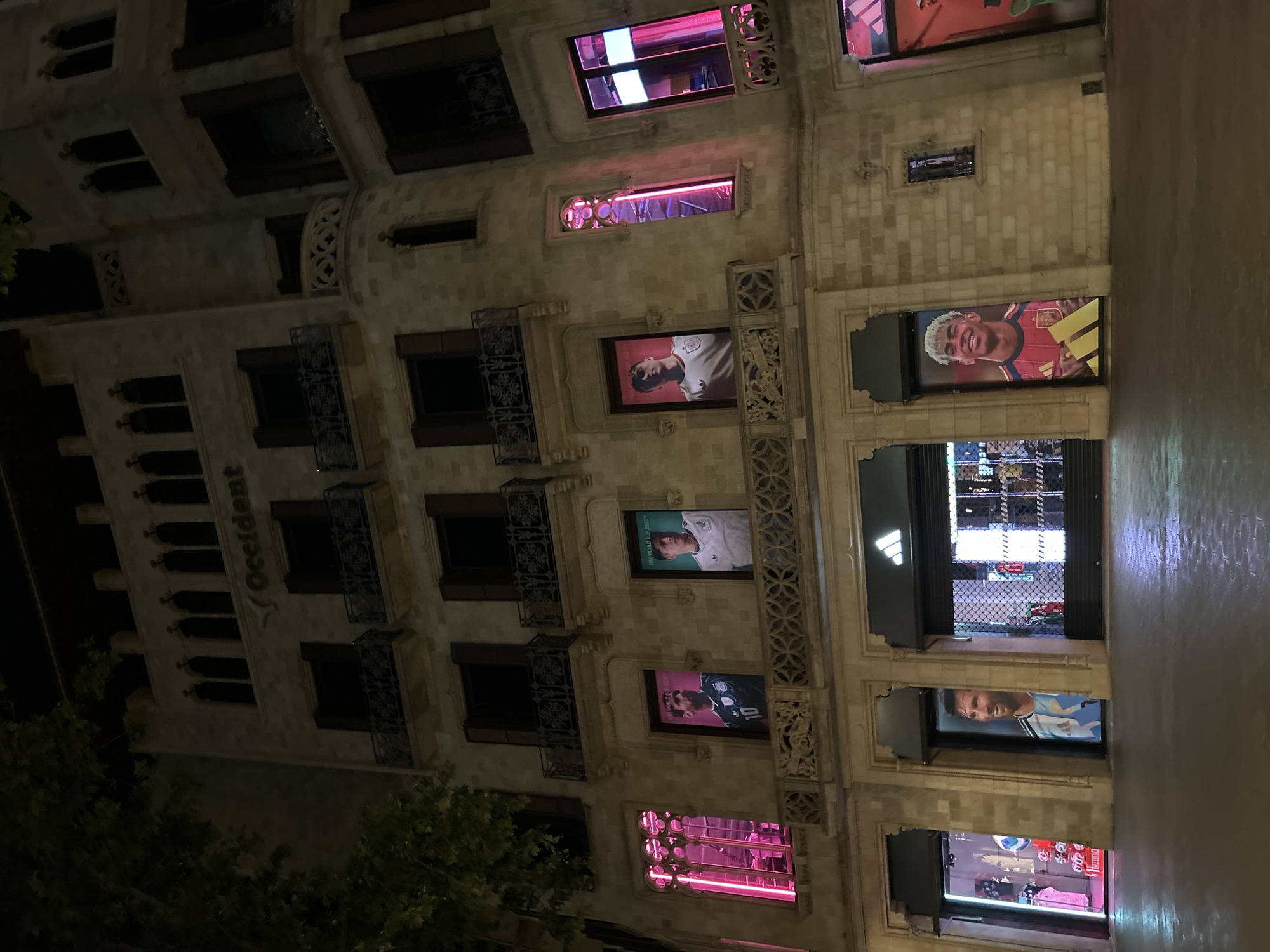 The image shows a tall, ornate building at night in Barcelona, Spain. The building, likely a commercial structure, has multiple stories with windows displaying sports-related advertising featuring soccer players. Pink neon lights are visible in some window displays. The street reflects ambient light, indicating recent or ongoing rain. Trees are partially visible on the lower left side of the frame. The word "Occident" is vertically displayed on the building's façade. The scene suggests an urban environment at night, likely in a tourist or commercial area.