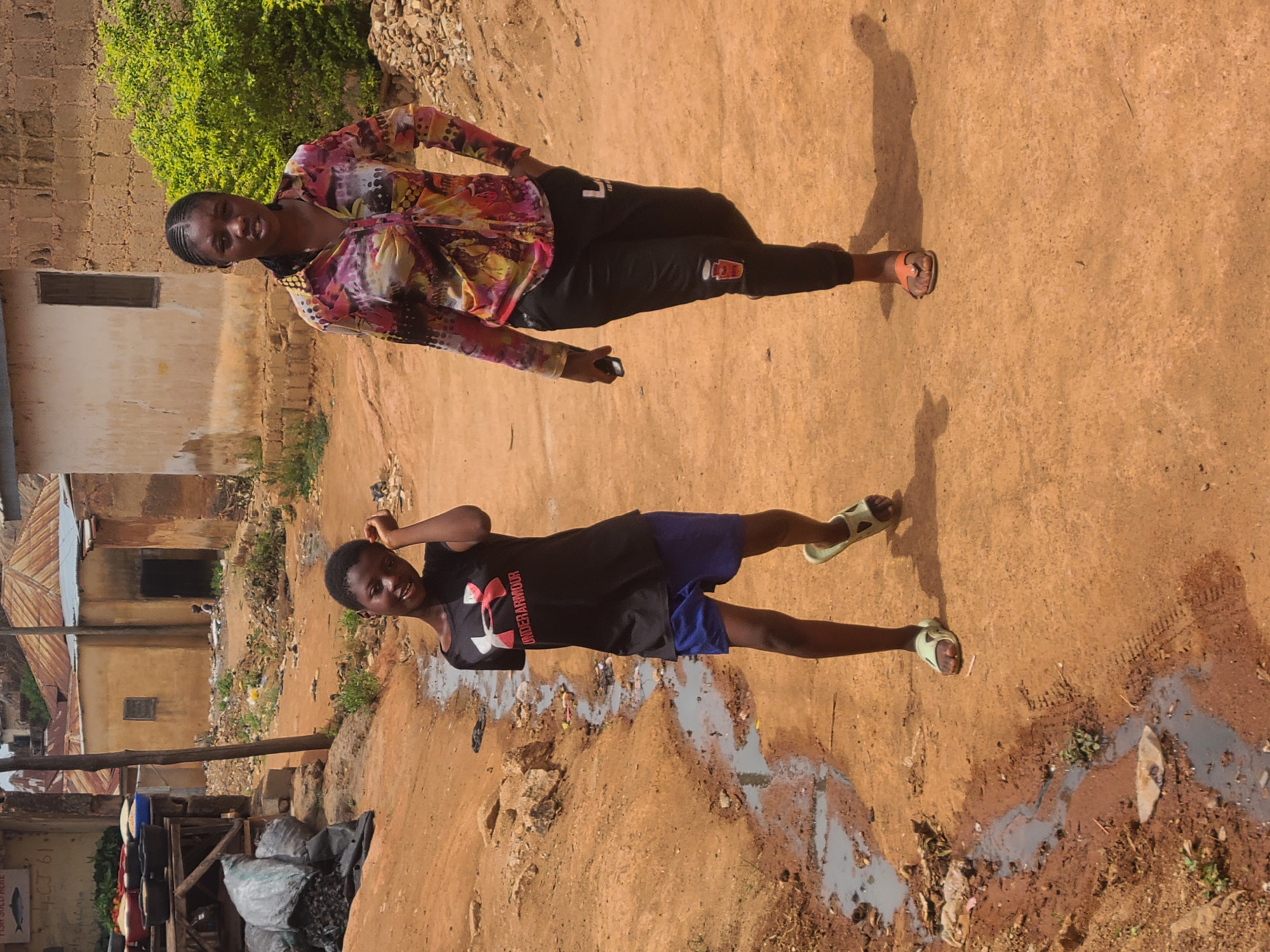 Two individuals are depicted in an outdoor setting. One is wearing a long-sleeved, multi-colored top with dark pants, while the other wears a dark t-shirt with blue shorts. The background features buildings, some with corrugated metal roofs, indicative of a developing urban area. The ground appears to be a dirt path, potentially in a lower-income or less developed section of Jos, Nigeria. There is some refuse and water on the ground. Based on the structures and environment, the location seems to be a residential neighborhood in the city.