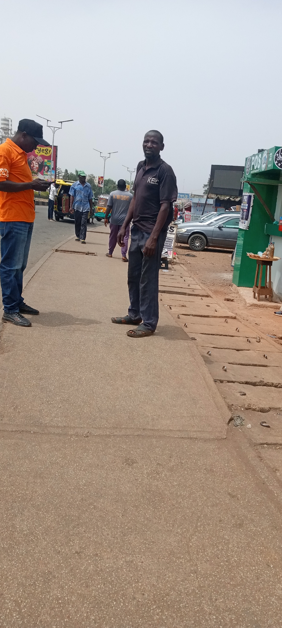 The image depicts a street scene in Jos, Nigeria. Two men are standing on a concrete sidewalk. The man on the left is wearing an orange shirt, blue jeans, and a black hat, and is looking at something in his hands. The man on the right is wearing a dark shirt with "KEC" visible on it, dark pants, and sandals, and is looking towards the camera. Behind them, several other people are walking along the sidewalk and the street. A yellow vehicle is visible in the street. To the right of the standing men is a green stall with "POS" written on it, and a small wooden table with items on it. The road is brown with some loose debris scattered on the side. Several metal protrusions are embedded in the concrete near the sidewalk. The sky is overcast.