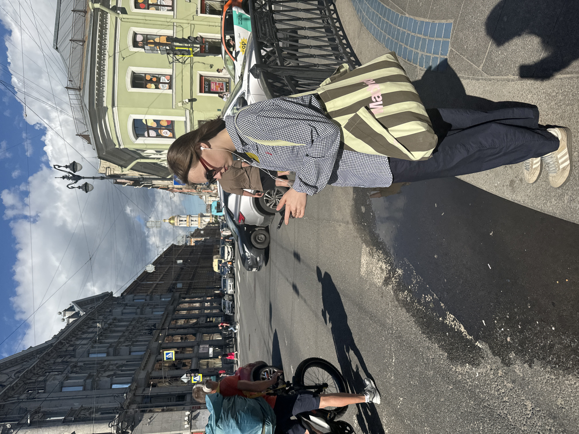 The image captures a street scene in Saint Petersburg, Russia. In the foreground, a young woman with sunglasses and white earbuds stands on a sidewalk, looking down at her phone. She is wearing a checkered long-sleeved shirt, dark pants, and light-colored sneakers. A striped tote bag hangs from her shoulder. Next to her, a man on a bicycle is also wearing earbuds and a large blue backpack. He is dressed in a red t-shirt and dark shorts, with grey sneakers. The bicycle has a large front wheel. The street is wet in places, suggesting recent rain. In the background, a variety of buildings line the street, with a golden-domed church visible in the distance. Cars are present on the road, and pedestrians are visible on the sidewalks. The sky is partly cloudy. The architecture suggests a historic urban setting.