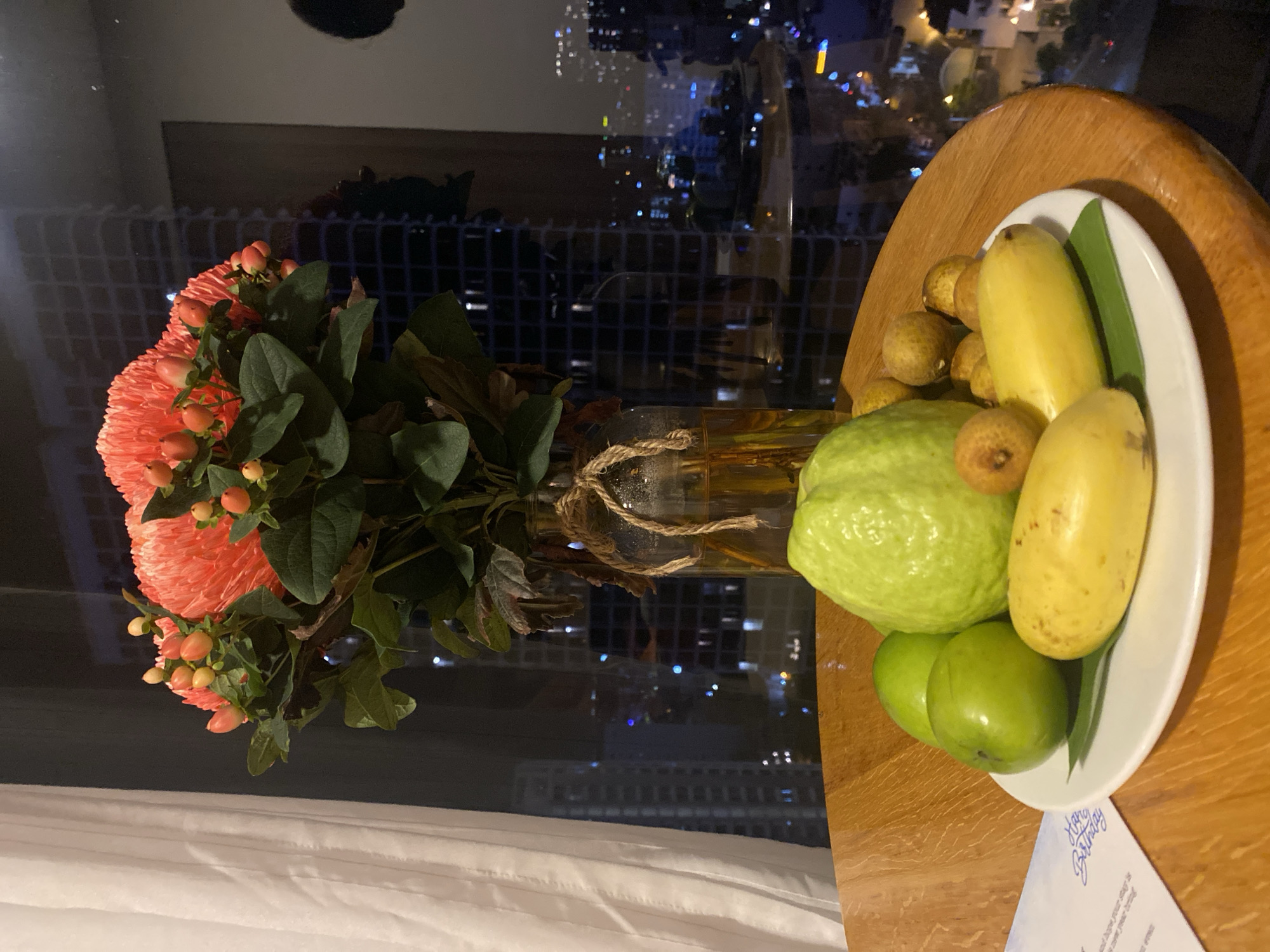 A still life arrangement of fruit sits on a wooden table in front of a window overlooking a city at night. The fruit includes a large, light green guava, several small, brown, round fruits (likely longan or lychee), two yellow bananas, and three small green apples. The fruit is arranged on a white plate lined with a green leaf. Behind the fruit is a glass vase containing a bouquet of coral-colored flowers with small, round, yellow buds. The vase is tied with twine. A white curtain hangs on the left side of the image. The window behind the arrangement reveals a cityscape with numerous illuminated buildings and lights, suggesting an urban setting, possibly in Nha Trang Ward, Vietnam. The lighting is dim, with artificial lights from the city providing illumination.