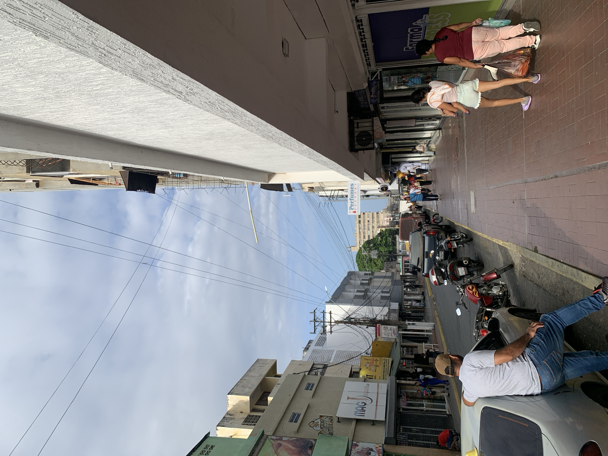 The image captures a vibrant daytime street scene in Porlamar, Venezuela, viewed from beneath a textured, light-colored building overhang. The sky above is partly cloudy.
On the right side of the frame, a brick-paved sidewalk extends into the distance, bustling with pedestrians. Two women are prominently featured in the foreground: one in a dark red top and light pink pants, carrying shopping bags, and another in a light-colored top and shorts, also with bags, walking ahead of her. Further down the sidewalk, more people are visible near storefronts.
Adjacent to the sidewalk is a street with multiple parked motorcycles along the curb. In the immediate foreground of the street, a man in a grey t-shirt, jeans, and a baseball cap is leaning casually against the rear of a silver car.
The left side of the image showcases a row of commercial and residential buildings with varied architectural styles and colors. A dense network of utility wires crisscrosses overhead, connecting to numerous power poles. Visible text on storefront signs includes "Farma" on a blue sign, "PERFUMES FACTORY" on a red and white sign, and "MAG import.ca" on a white sign, indicating various businesses. The scene depicts a lively urban environment, typical of a commercial district.
