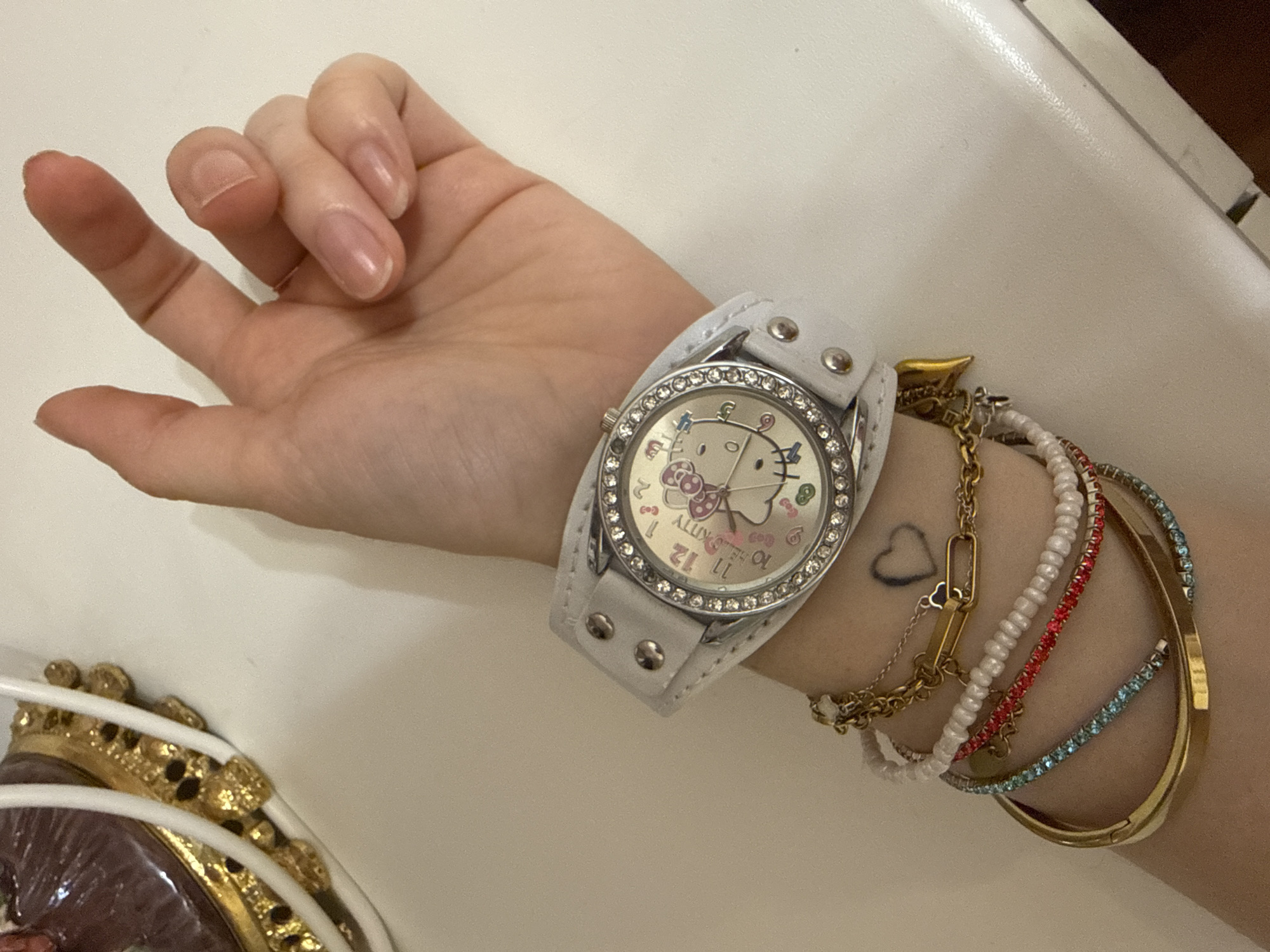 A close-up shot shows a person's left wrist adorned with a white watch featuring a Hello Kitty face and a crystal-encrusted bezel. Several bracelets are stacked on the wrist below the watch: a gold chain bracelet, a white beaded bracelet, a bracelet with red rhinestones, a bracelet with blue rhinestones, and a thick gold bangle. A faint heart tattoo is visible on the wrist. The background is a plain white surface, suggesting an indoor, possibly domestic setting.