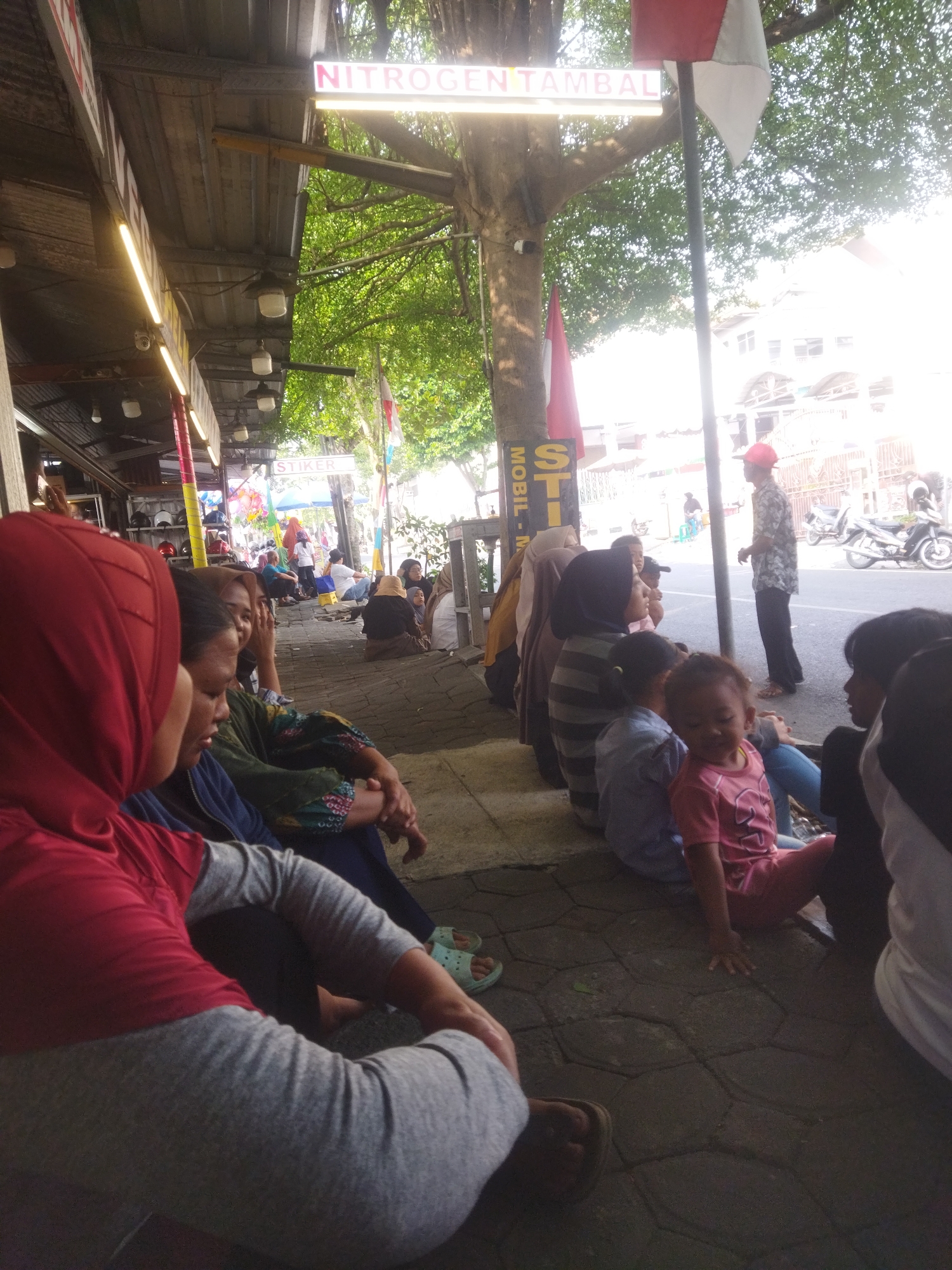The image shows a group of people sitting on a sidewalk in front of a covered market stall. A sign above the stall reads "NITROGEN TAMBAL". The people appear to be waiting or resting. In the background, there are buildings, trees, and a street with motorcycles. The scene suggests a commercial area in a city, likely in Indonesia, given the context of the sign and the appearance of the people and surroundings. The lighting suggests it is daytime.