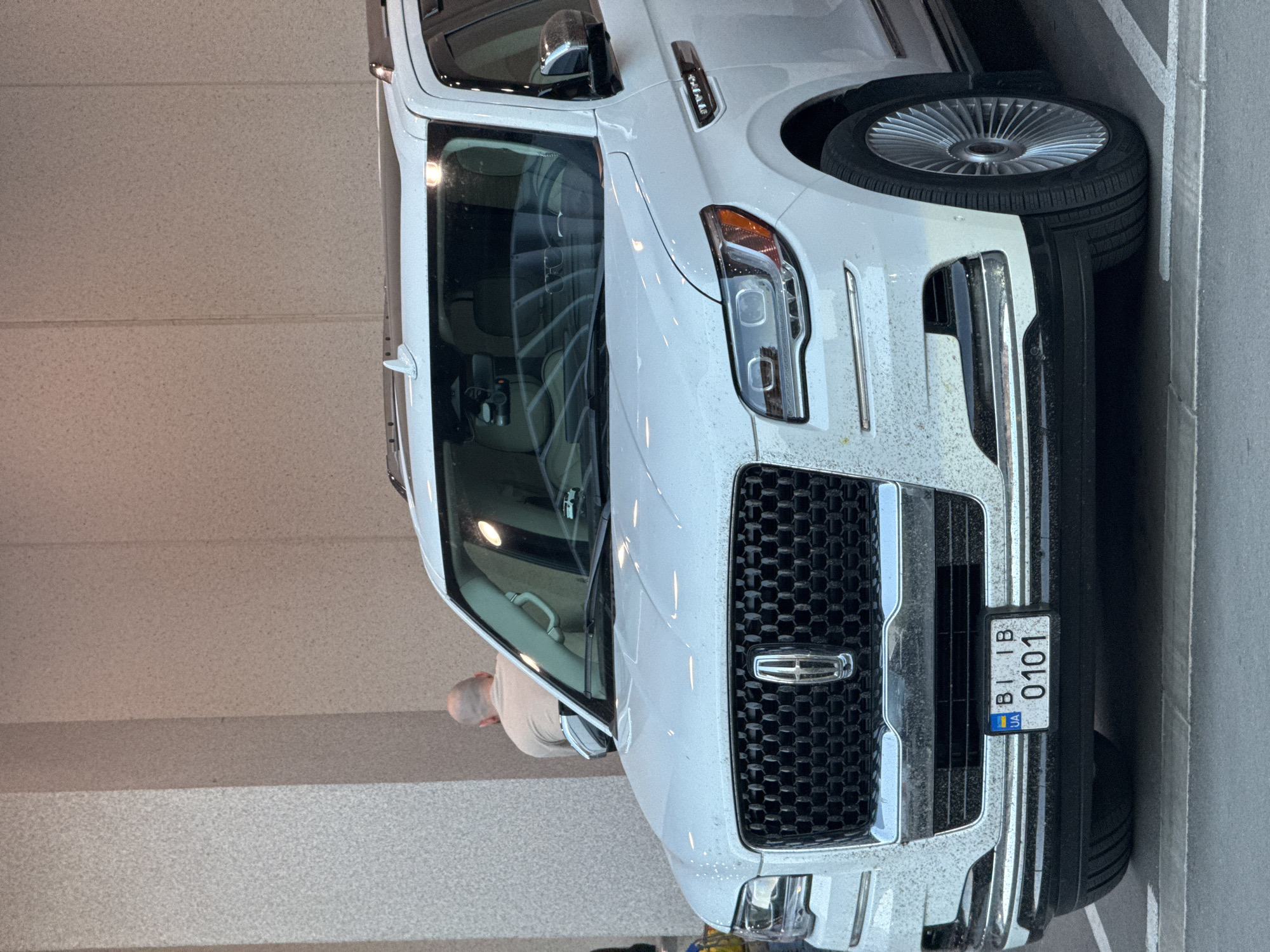 A white Lincoln Navigator SUV is parked in what appears to be an outdoor parking area with textured light-colored walls in the background. The vehicle's front grille, headlights, and license plate are clearly visible. The license plate is from Ukraine, reading "BI 1B" above "0101". A person with a shaved head is partially visible in the background, wearing a light-colored t-shirt and facing away from the camera, near the entrance of a building. The SUV has visible water spots or dirt on its exterior. The ground is paved with what looks like concrete or asphalt, marked with white parking lines. The scene is captured in daylight.