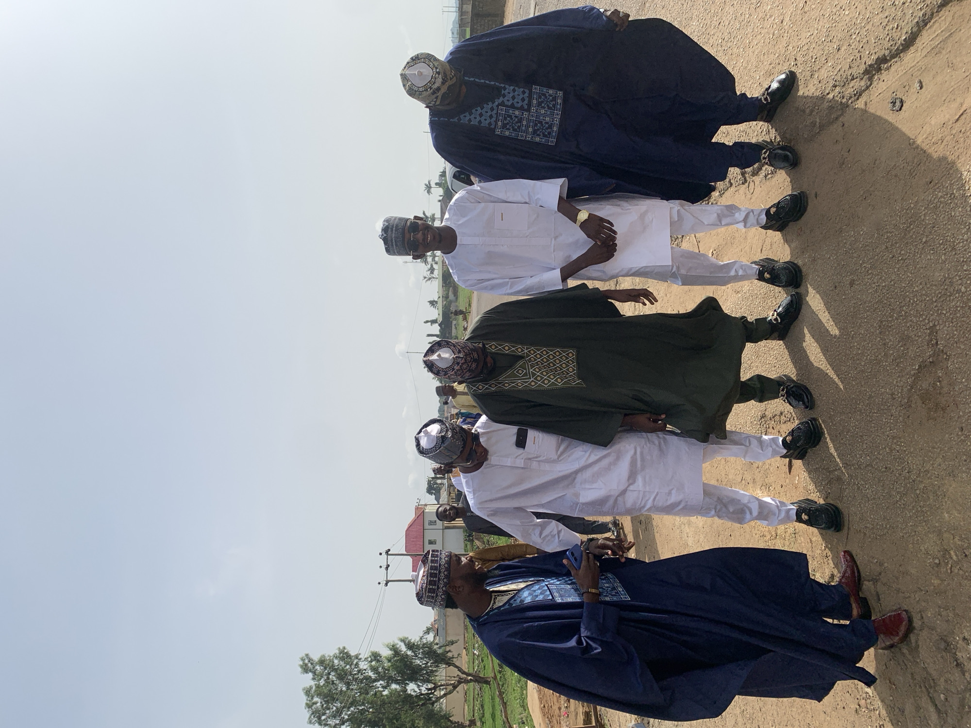 Five men are standing on a dirt road. All are wearing traditional Nigerian garments and caps. The man nearest the camera is wearing a dark blue robe with detailed embroidery and maroon shoes and is speaking on a cell phone. The next two men are wearing white robes. The fourth man is wearing a dark green robe with gold embroidery. The man farthest away is wearing a dark blue robe. The background contains buildings, trees, and utility poles. The location is Naraguta Hausawa, Nigeria.