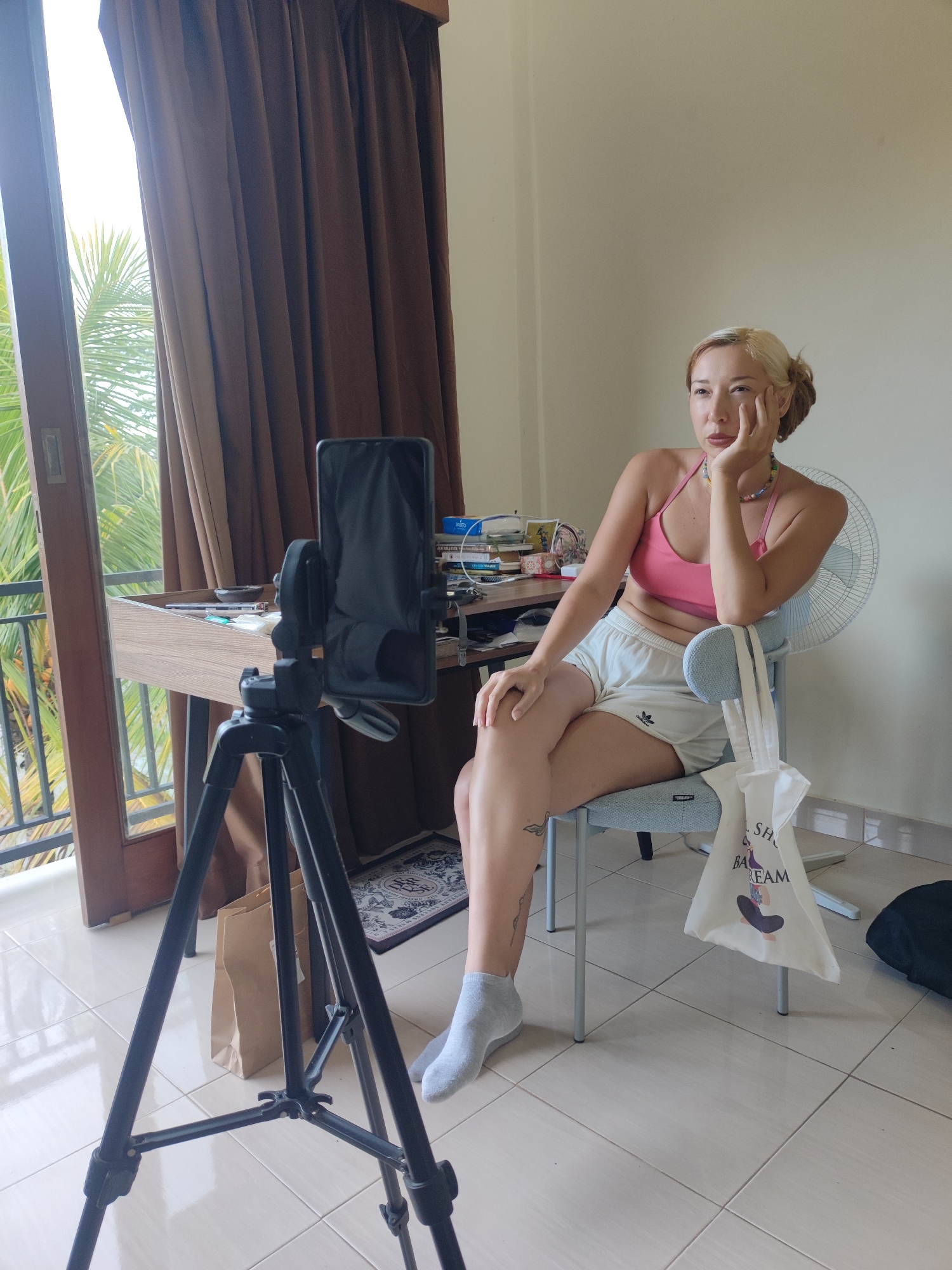A woman with bleached blonde hair and fair skin sits in a light blue office chair in a well-lit room, likely in Tegallalang, Indonesia. She wears a pink halter top and white Adidas shorts. A single grey sock covers her left foot. Her left elbow rests on the back of the chair and her head is supported by her left hand. A white tote bag hangs from the back of the chair. In front of her is a black smartphone attached to a tripod, likely recording her. A desk cluttered with various items stands behind the phone. Behind the desk, a large window shows a view of palm trees outside. Brown curtains hang to the right of the window. The floor is tiled in a light color.