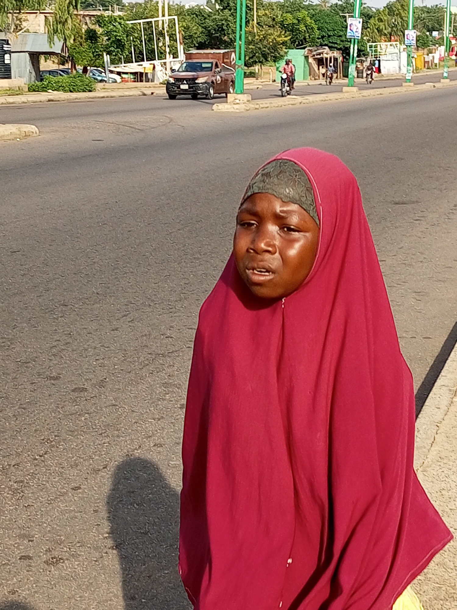 A young girl, appearing to be of African descent, stands facing left on a paved road. She wears a dark patterned headscarf and a bright pink hijab that covers her head and shoulders. Her mouth is slightly open, and her expression suggests she might be speaking or crying. Sunlight reflects off her face, highlighting her features. Her shadow is cast on the asphalt beside her. In the background, the road continues, with a curb and sidewalk visible on the right. Further in the distance, across the road, are various structures, including buildings with corrugated metal roofs, a pickup truck, and several people on motorcycles. Trees and poles with banners are also present, suggesting an urban or semi-urban environment. The overall lighting indicates it is daytime, likely under direct sunlight.