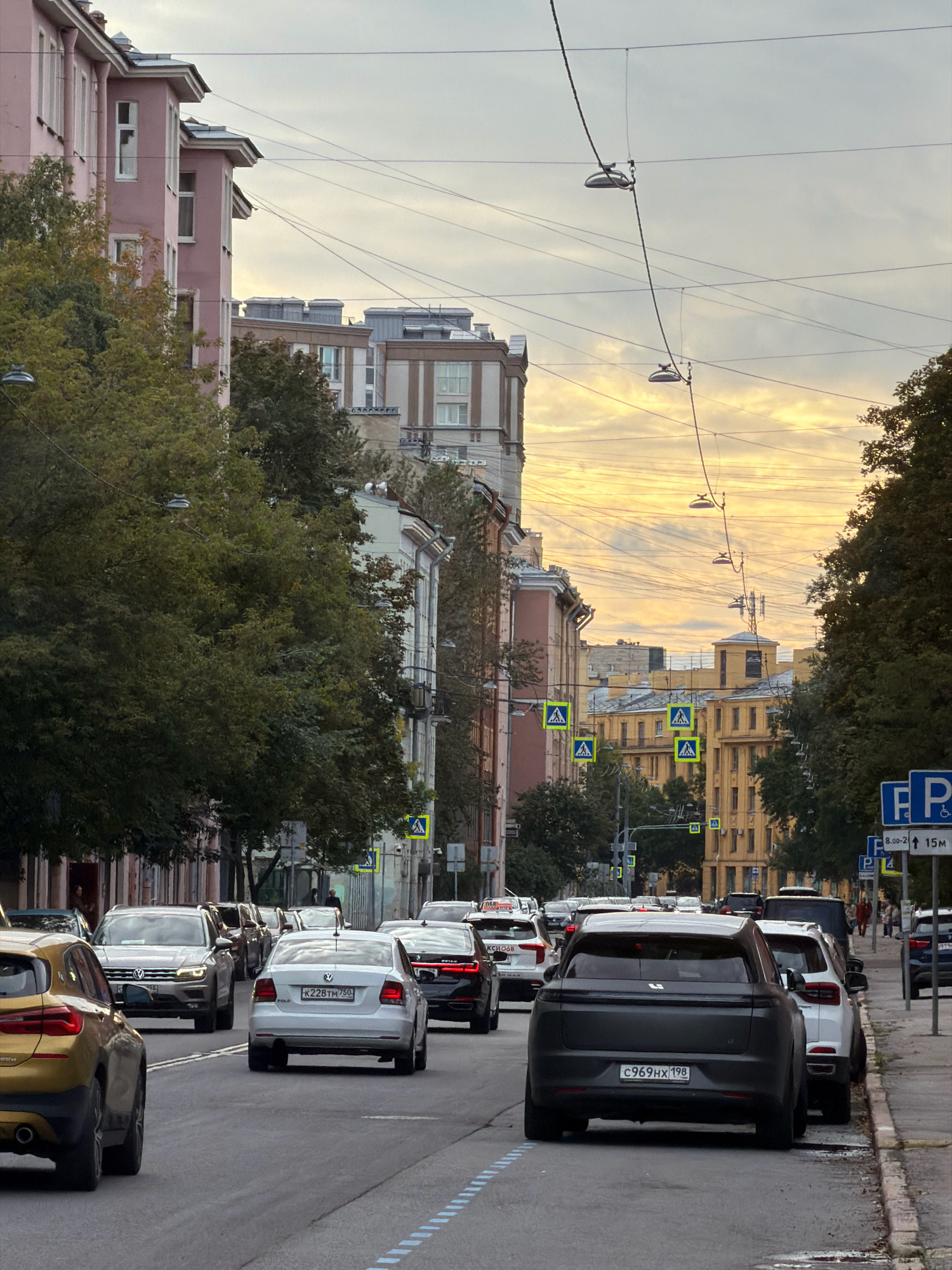 This image depicts a street scene in Saint Petersburg, Russia, likely during the late afternoon or early evening, given the warm, overcast sky. The perspective is from the middle of a street, looking forward, with traffic moving away from the viewer.
On the left side of the street, there are multi-story buildings with pink and beige facades, featuring numerous windows. Dense green trees with some autumnal foliage line the sidewalk in front of these buildings. Overhead, a complex network of electrical wires and streetlights is visible against the sky.
The street itself is paved, with visible dashed blue markings on the right lane. Several cars are driving or parked on the street. Prominent vehicles include a gold SUV in the foreground on the left, a white Volkswagen Polo with the license plate K228TH 750 in the center, and a dark-colored SUV with the license plate C969HX 198 closer to the right side of the frame. Other vehicles, including sedans and SUVs, are present in the traffic flow and parked along the right curb.
On the right side of the street, there are more buildings, some with yellow facades. Pedestrian crossing signs with yellow triangular backgrounds are visible at intervals along the road. Further down the street, a blue parking sign with a "P" symbol and parking time restrictions is mounted on a pole. A few pedestrians are visible in the distance on the sidewalk. The overall impression is of a typical urban street in a city with a mix of residential and possibly commercial buildings.