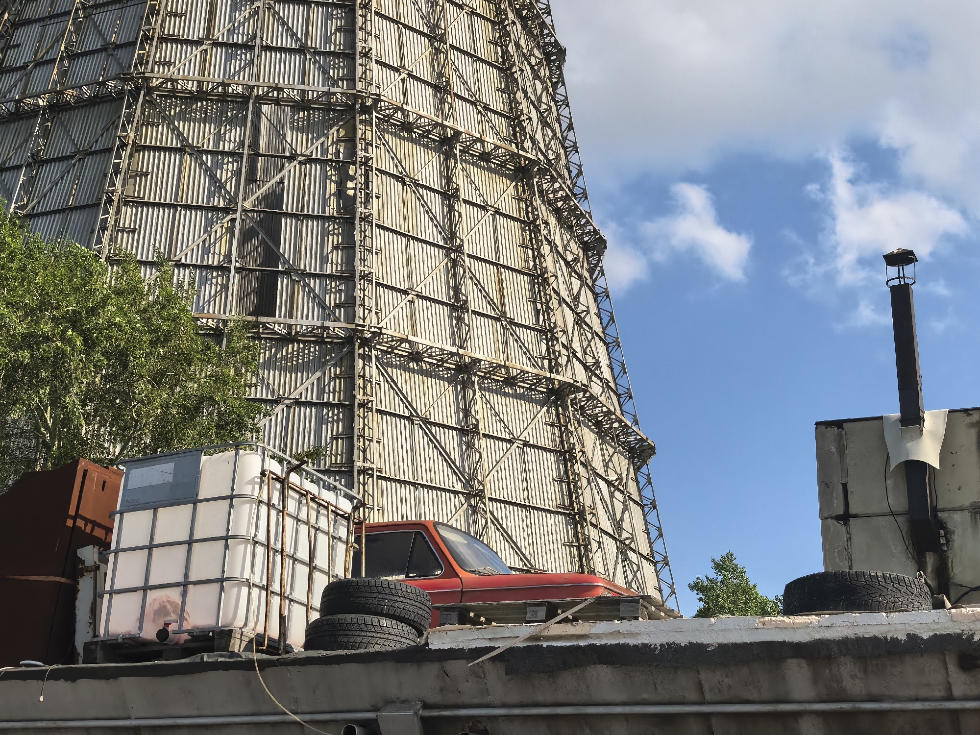 In an industrial outdoor setting, a large, cylindrical structure made of metal scaffolding and corrugated panels dominates the background. In the foreground, a red car is partially visible, parked on a raised concrete platform. Next to the car, there is a large white plastic tank with a metal cage. Several car tires are stacked on the platform, and another tire sits on a separate structure to the right. A dark chimney extends upwards on the right side of the image, against a blue sky with scattered white clouds. Green trees are visible behind the car and tank. The overall scene suggests an abandoned or disused industrial area.
