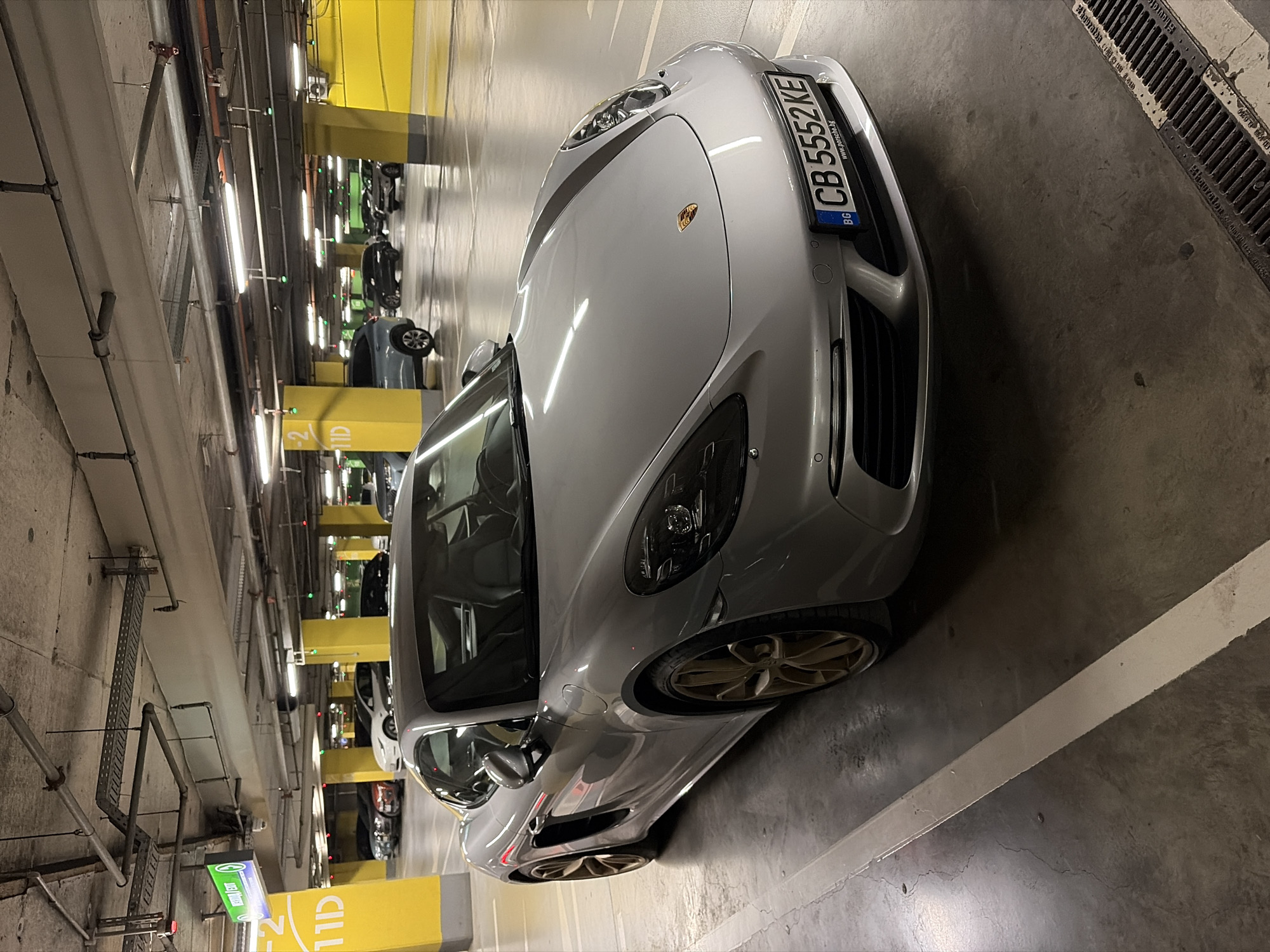 A silver Porsche sports car is parked in an underground parking garage. The car is facing towards the right of the frame, with its front quarter visible. The license plate reads "CB 5552 KE" and has "BG" indicating Bulgaria. The car is parked between white lines on a concrete floor, with a drain grate visible in the lower right corner. In the background, other cars are parked, and yellow pillars and concrete ceilings with exposed pipes and lighting fixtures are visible. A green exit sign with Hebrew text is visible in the upper left quadrant. The scene appears to be in a parking garage in Sofia, Bulgaria, as indicated by the license plate and the Hebrew text on the sign which is likely a mistaken interpretation of Cyrillic letters often used in Bulgarian signage, or a multilingual sign in a location frequented by tourists.