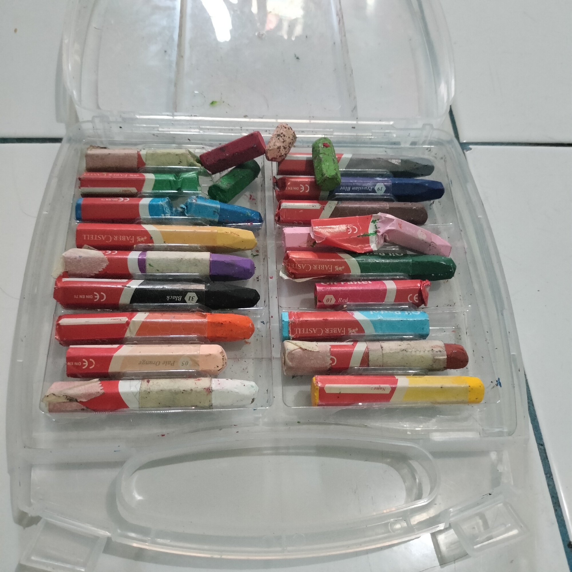 An open, clear plastic container holds a set of oil pastels. The container is placed on a white tiled surface. The pastels are arranged in two columns, with some scattered on top. The pastels are individually wrapped in paper, with some wrappers showing brand names and color information. The container itself appears to have some dust and debris inside. The scene is indoors, with overhead lighting reflecting on the plastic lid of the container. There are no people or animals visible in the image. The location in Tangerang, Indonesia is not specified within the image.