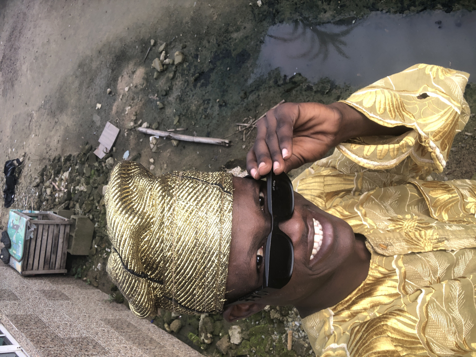A man, wearing a gold cap and a gold patterned shirt, is taking a selfie. He is wearing black sunglasses and is smiling. Behind him, there is a body of dirty water with some debris. To his left, there is a wall with a wooden structure on top of it. The environment appears to be an outdoor area in Ado, Nigeria.