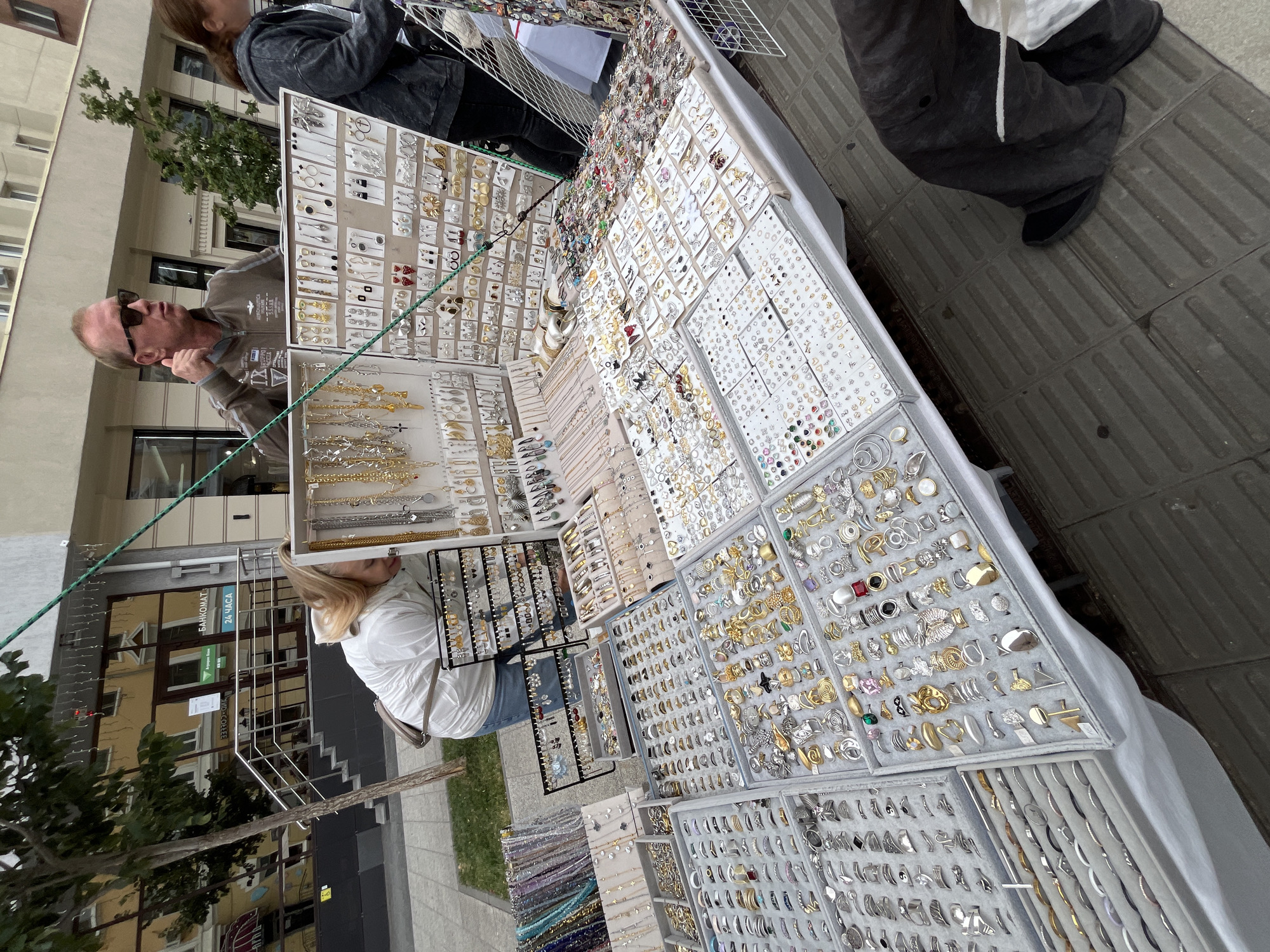 The image depicts an outdoor jewelry market, likely in Saratov, Russia, as indicated by a sign in the background that reads "БАНКОМАТ" (ATM) and "24 ЧАСА" (24 hours), common phrases in Russian. A woman with blonde hair, wearing a white jacket and blue jeans, is seen bending over a display table filled with jewelry, examining the items. In the background, a man wearing sunglasses and a brown jacket gives a thumbs-up gesture. The market features numerous tables laden with a wide assortment of jewelry, including necklaces, earrings, and rings, displayed on white fabric and in organized trays. The backdrop includes a building with windows reflecting the street, a tree, and a paved walkway. The overall scene captures a commercial exchange environment with a variety of jewelry for sale.
