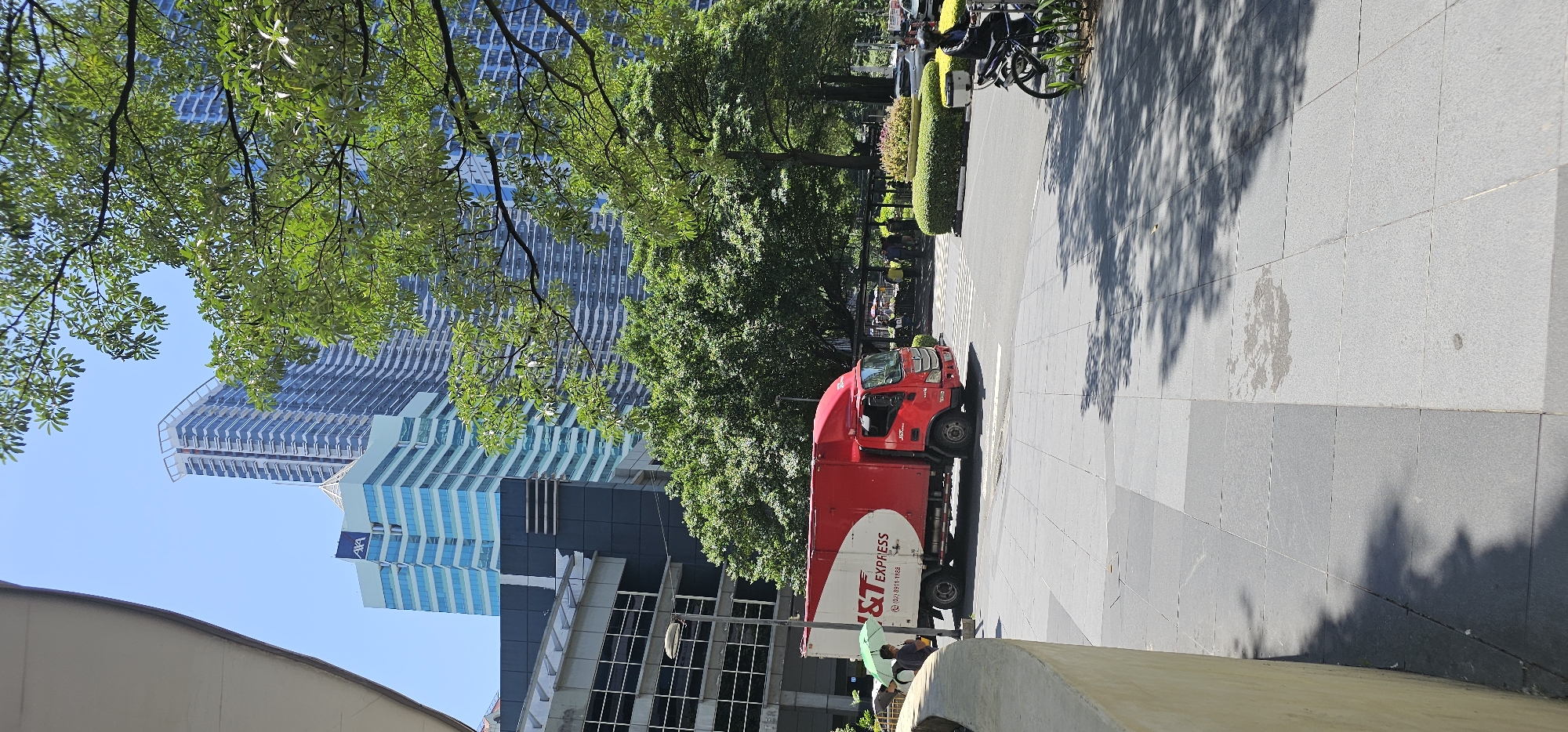 This image depicts a street scene in Makati, Philippines. In the foreground, a red and white J&T Express delivery truck is parked on the right side of a paved road. The truck has a white cargo box with a red stripe and the J&T Express logo. To the left of the truck, a person holding a light green umbrella stands near a white vehicle. Further to the left, a portion of a curved, light-colored wall is visible.
In the background, several modern buildings with glass facades rise against a clear blue sky. One building has the "AXA" logo at its top. Lush green trees are scattered throughout the scene, with dense foliage obscuring some of the buildings. A motorcycle with a rider is parked on the right side of the frame, near a well-maintained, circular bush. In the distance, under a covered area, several people are visible, some seated. The ground is paved with large, gray tiles. Sunlight casts dappled shadows across the pavement.