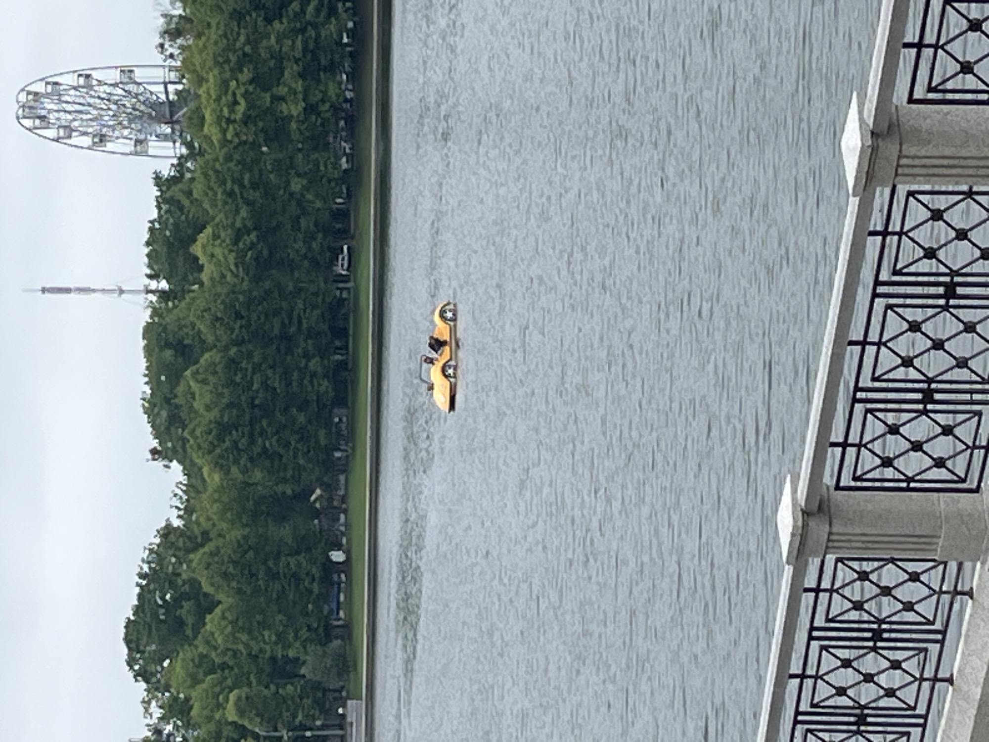 An orange pedal boat shaped like a car is in the middle of a body of water. Two people are visible in the boat, one appearing to steer. In the background, a Ferris wheel and a tall antenna are visible behind a line of green trees. A decorative fence with stone posts runs along the bottom of the image, framing the water. The sky is overcast. This scene is likely at the Upper Pond in Kaliningrad, Russia, given the presence of the Ferris wheel, which is a notable landmark at the park surrounding the pond.