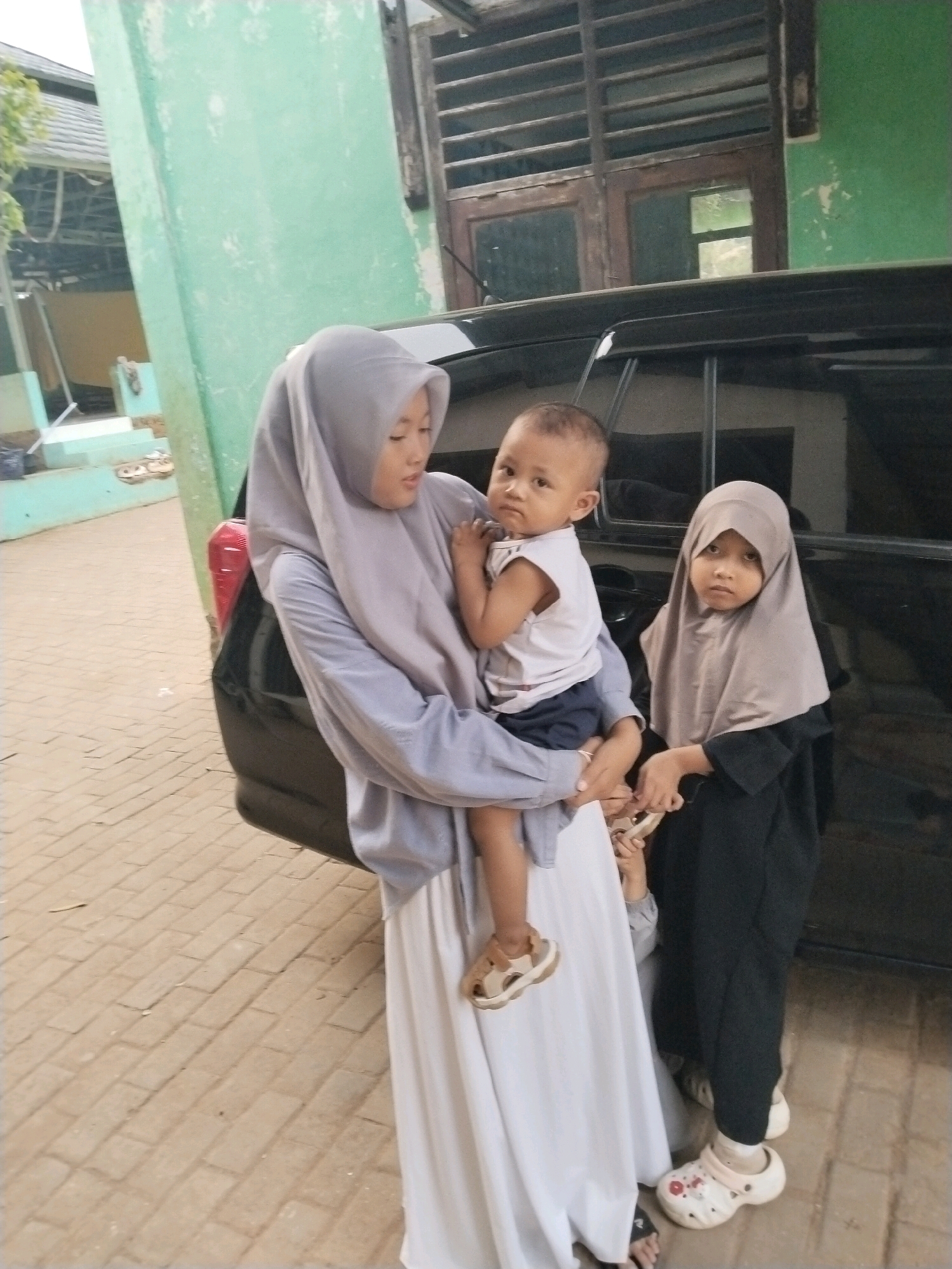 In Serang, Indonesia, three young children are positioned near a dark-colored vehicle. The child on the left, appearing to be a girl wearing a light gray hijab and a long white dress, is holding a baby boy. The baby boy is dressed in a white t-shirt and dark shorts. To the right of the girl, a younger child, also appearing to be a girl wearing a beige hijab and a dark abaya, stands by the vehicle. The ground beneath them is paved with light brown rectangular bricks. In the background, a light green wall of a building is visible, with a section of wooden-slatted windows.