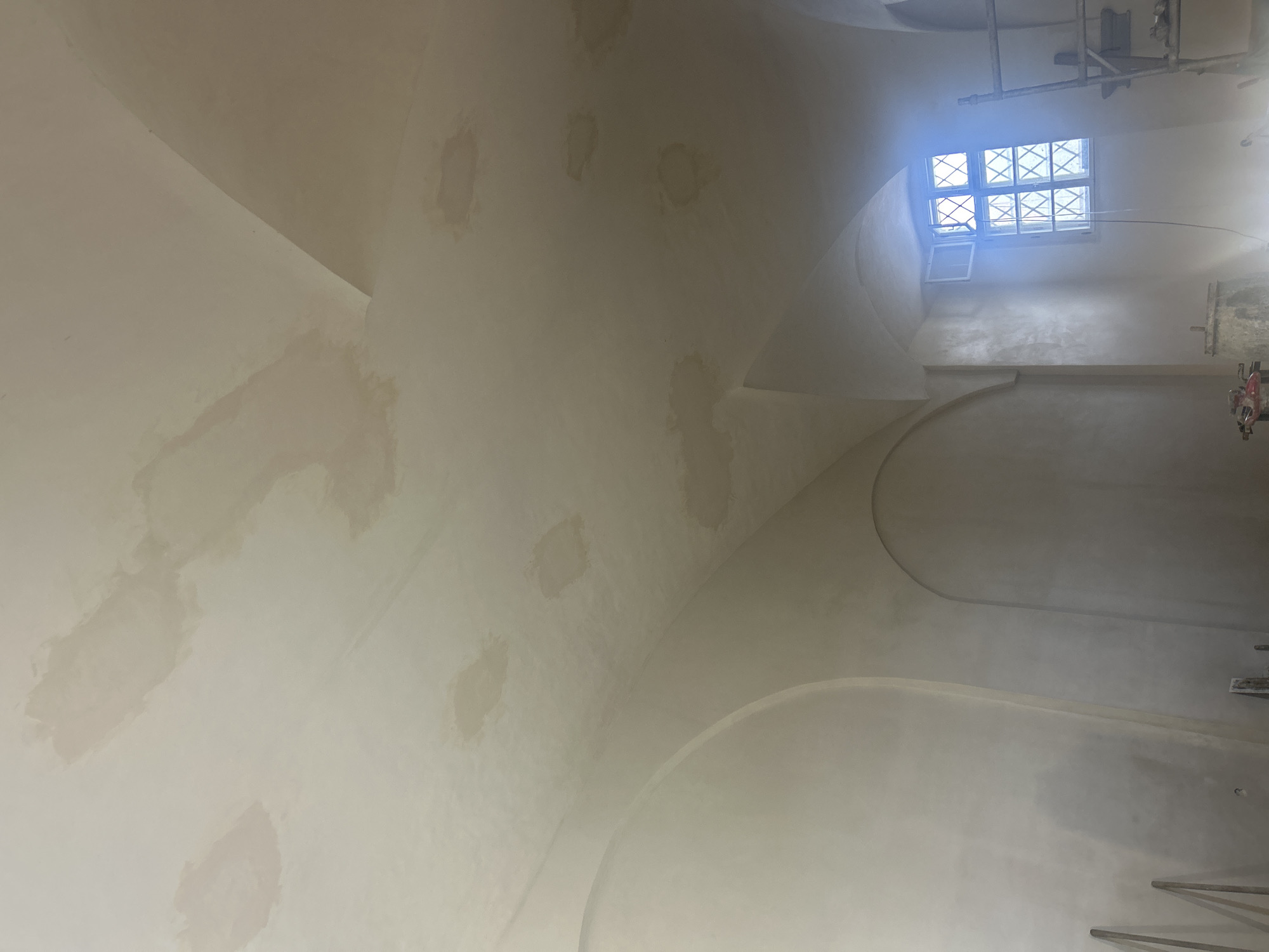 This is a close-up, eye-level, interior shot of a ceiling in what appears to be a historic building undergoing renovation, possibly located in Rosice, Czechia. The ceiling is made of a pale, slightly textured material and shows numerous splotchy water stains in various shades of beige and light brown, suggesting previous water damage. These stains are unevenly distributed across the arched sections of the ceiling. In the background, a window with a diamond-shaped grille is visible, and a portion of a scaffolding structure is present on the right side of the frame. The lighting is somewhat diffuse, with a bright glare emanating from the window area. The overall impression is one of an unfinished space with visible signs of repair and restoration work.