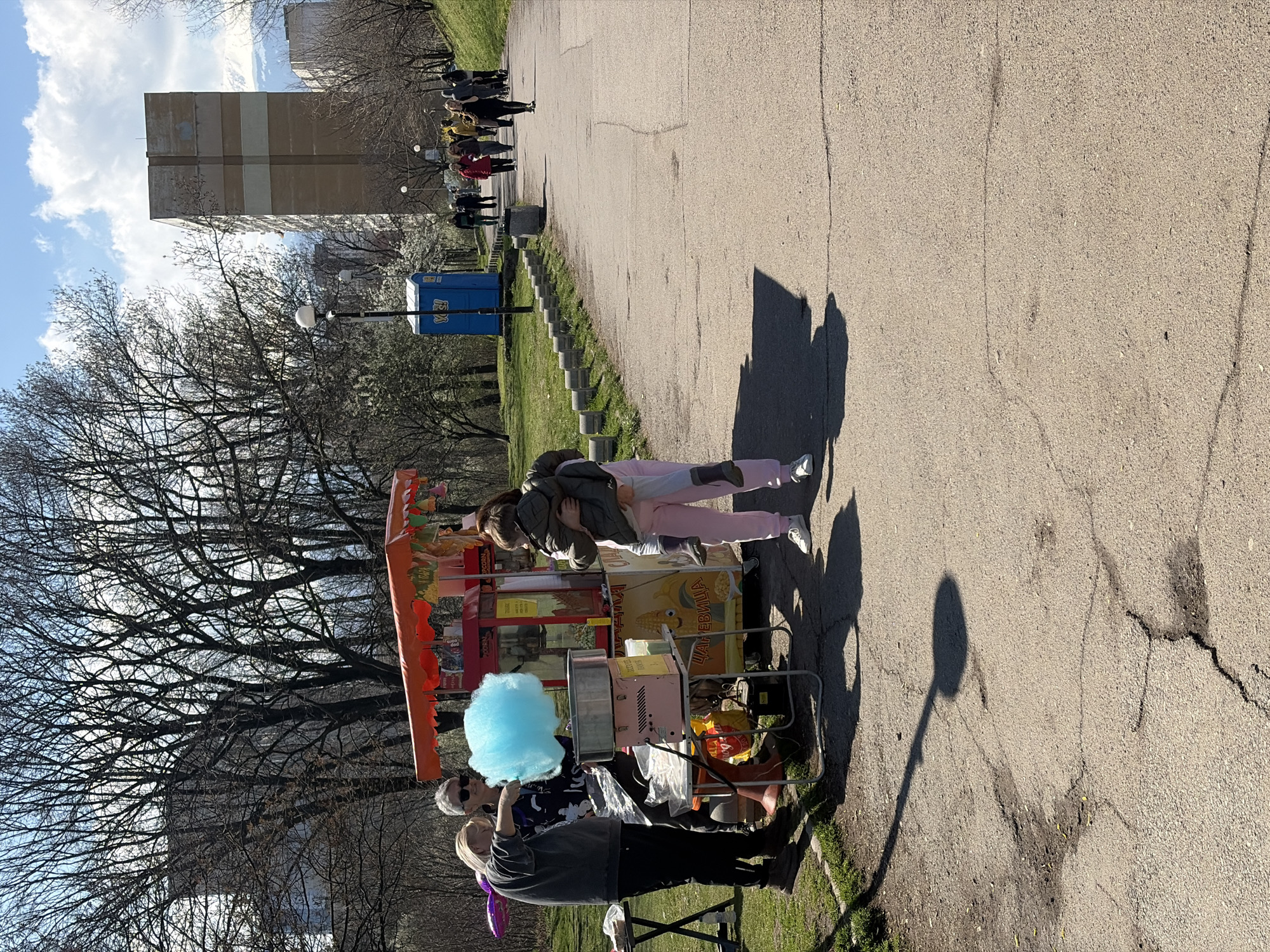 The image shows an outdoor scene in what appears to be a park. In the foreground, there is a vendor with a cotton candy cart. The cart is colorful and loaded with treats. Two people are at the cart, seemingly purchasing or interacting with the vendor. One appears to be purchasing cotton candy, as the vendor is making it. The cotton candy is light blue. A young girl and an adult are hugging and facing the cart. Further back, a line of people can be seen walking along a path, perhaps also enjoying the park. In the background, there is a large apartment building, the top half of which is obscured by a cloudy sky. There is a public toilet in the distance to the right of the apartment building. The ground is a paved path. The presence of Cyrillic text on the cart implies that the location is in a country that uses the Cyrillic alphabet, such as Bulgaria. Given the presence of the apartment building in the background and the type of trees, it is likely that this photo was taken in the city of Sofia, Bulgaria.