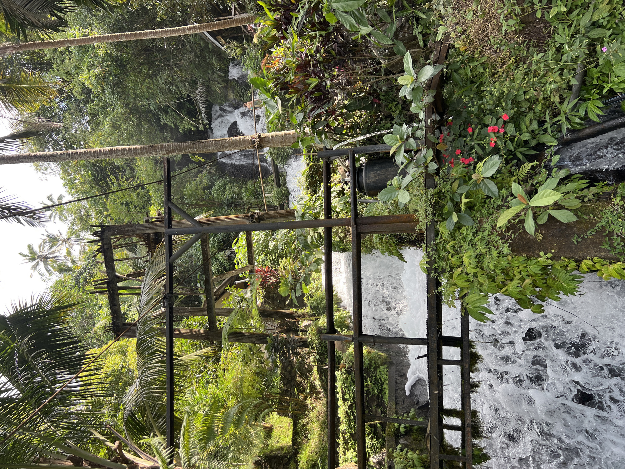 The image shows a lush, verdant natural scene in Sukasada, Indonesia, featuring a multi-tiered waterfall cascading down a rocky incline. In the foreground, a rustic wooden structure with metal beams appears to be part of an old water management system, possibly an old irrigation or milling mechanism, with water flowing through channels and over wooden planks. The surrounding environment is dense with tropical vegetation, including palm trees, ferns, and various flowering plants with red and pink blossoms. In the background, the waterfall is visible, with two individuals bathing or standing near the water's edge. The overall impression is one of a serene, natural setting with evidence of human intervention in the form of the wooden structure.