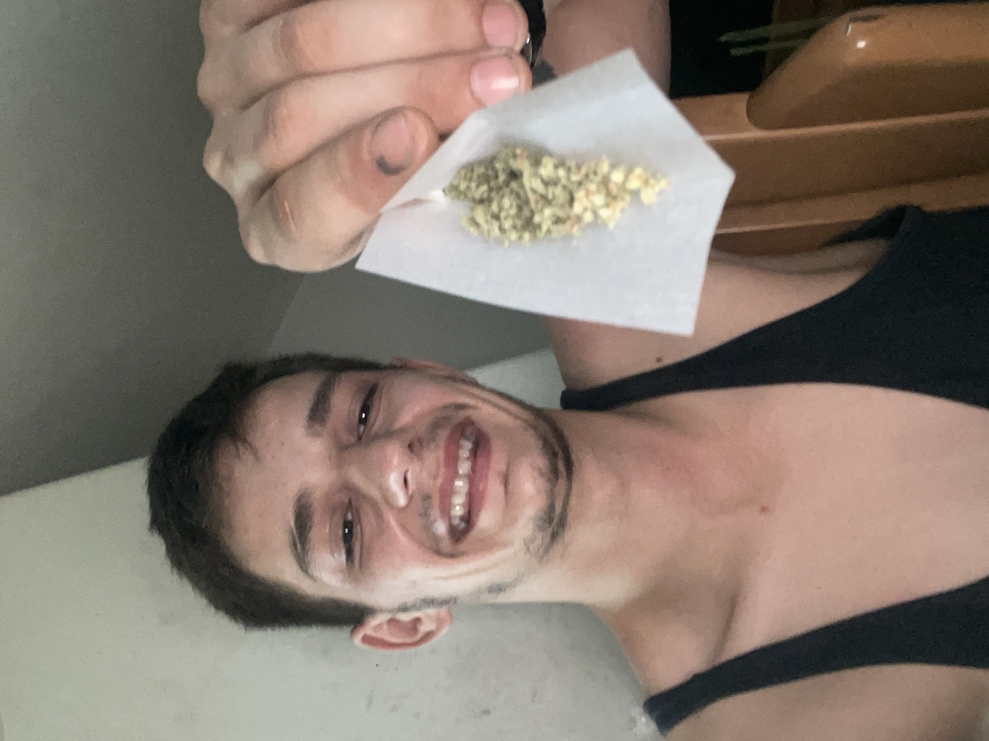A young adult male, with short brown hair and a thin build, is holding a rolled marijuana cigarette in his right hand towards the camera. He is wearing a black tank top and has a dark mole on his neck. The background is a plain, light-colored wall and a portion of a brown wooden object. The lighting is dim, and the image appears to be a selfie.