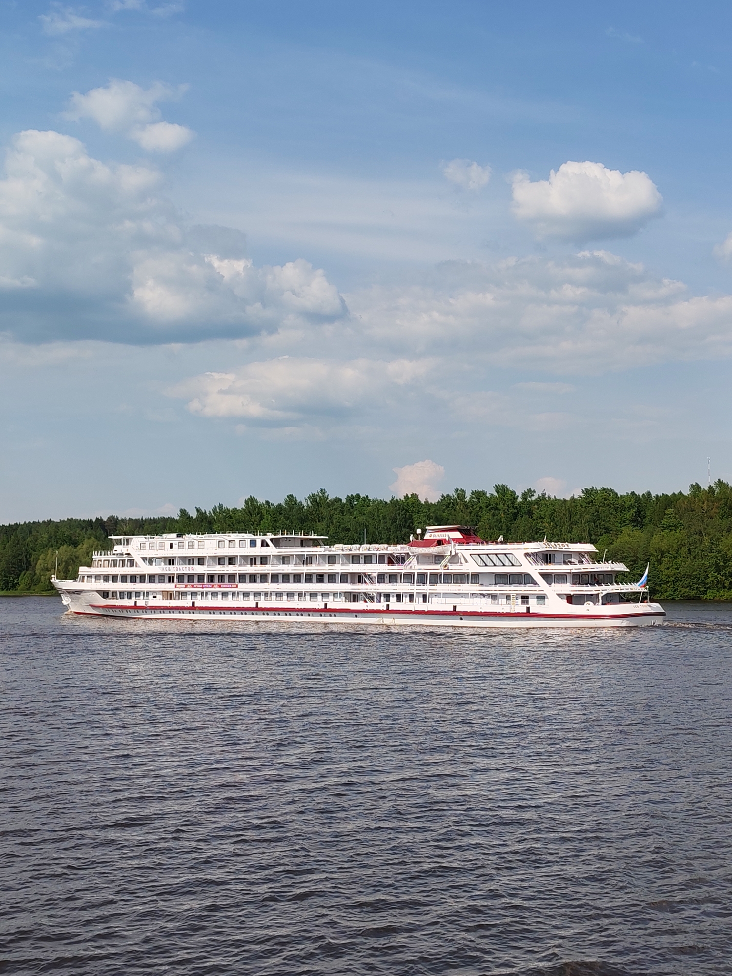 A white passenger ship with red accents is moving on a body of water, possibly a river or lake. The ship has multiple decks with windows and appears to be several stories high. Behind the ship is a line of green trees, indicating the shoreline. The sky is blue with scattered white clouds. The water is dark with slight ripples. The ship is the "Lev Tolstoy," a four-deck motor ship of the 302 project, built in Germany and renovated in 2017. It is located on the Moscow Canal near the village of Trudovaya, Russia. There are no people visible in the image.