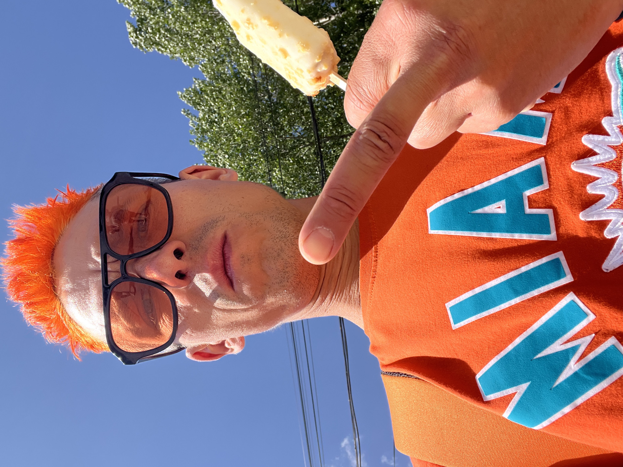 A low-angle shot shows a person with orange hair and black sunglasses, wearing an orange Miami Dolphins sweatshirt, pointing at a popsicle. The background features a blue sky, a tree, and overhead power lines. The location is not discernible from the image.