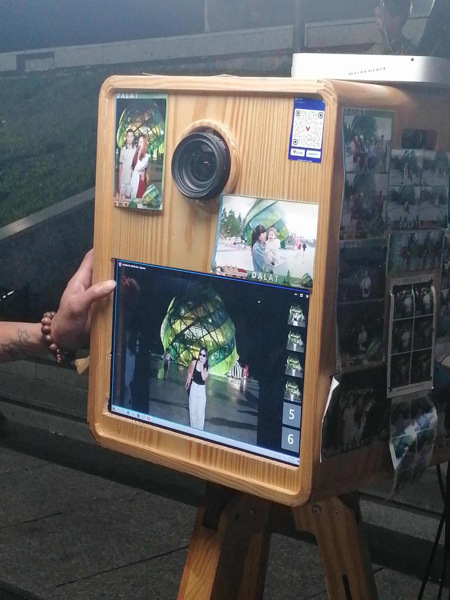 A photo booth crafted from light-colored wood stands outdoors on a paved surface, likely in the city of Da Lat, Vietnam, as indicated by the printed images on the booth's surface. The booth features a dark-lensed camera and a computer screen displaying a live video feed showing a woman in a black tank top and white pants posing in front of a geometrically-designed glass building. To the left of the screen, a hand is visible with a bracelet. Various printed photos are displayed on the booth, including tourist scenes and couples. A white object sits on top of the booth. The booth has wooden legs.
