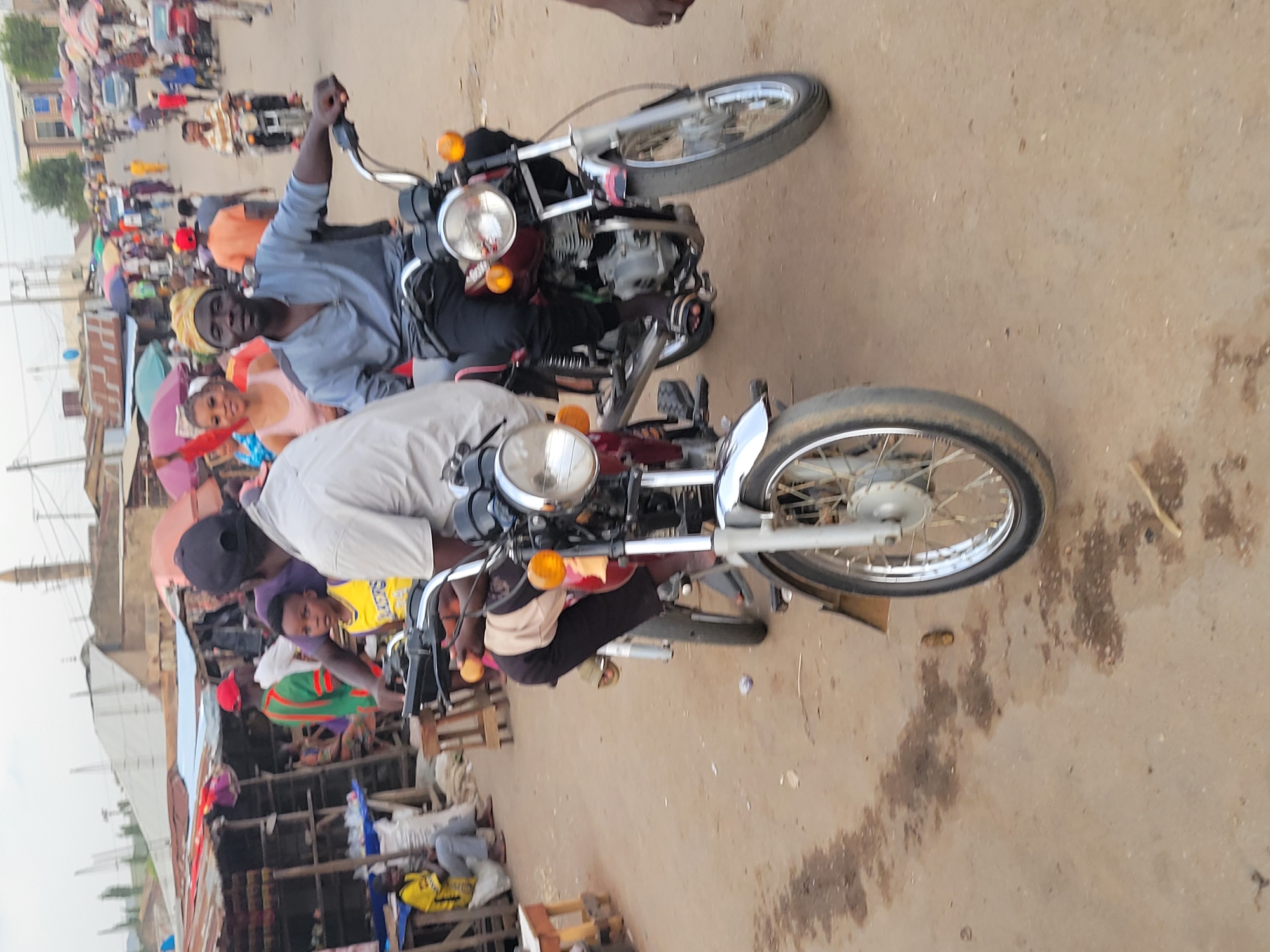 The image shows two men riding motorcycles on a street in Madalla, Nigeria. The street appears to be bustling with people and activity. There are numerous individuals on foot, some under umbrellas, suggesting a market or busy commercial area. Along the side of the road are makeshift stalls and structures, indicative of street vending. The motorcycles are simple, standard models, and the riders are casually dressed. The overall environment suggests a lively and possibly congested urban setting.