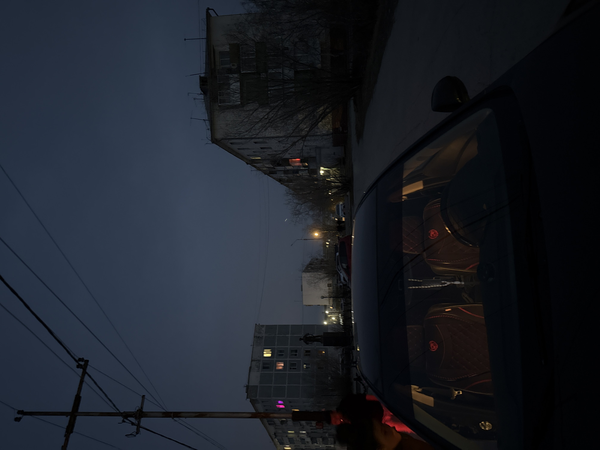 The image shows a nighttime street scene in Zhatay, Russia. Buildings line the street, lit by streetlights and a few interior lights. A vehicle is parked on the street, obscuring some of the view, with objects visible inside the car. Electrical wires are visible in the sky. A statue is visible, potentially of Lenin or another figure associated with the Soviet era.