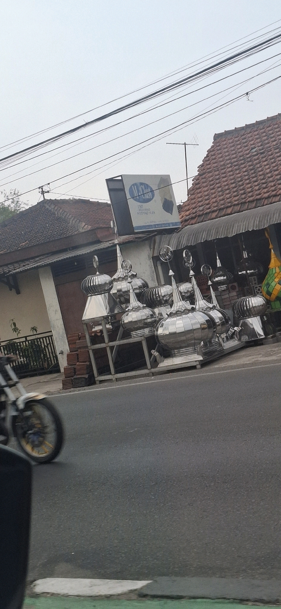 In the city of Ngadirejo, Indonesia, on a street with power lines overhead and a tiled roof building in the background, various architectural elements are displayed. In the foreground, a motorcycle is partially visible, its wheel blurred from motion. Directly behind the motorcycle and to the right, a collection of polished metal domes, likely intended for building ornamentation or ventilation, are arranged on a metal frame. These domes vary in size and shape, featuring pointed tops and ribbed surfaces. To the left of the domes, a stack of reddish-brown tiles rests on the ground. Above the domes and the building, a sign for "M.J Class" is mounted, advertising "THE PREMIUM ROOFING TILES". A portion of a green and yellow decorative object is visible on the far right. The overall scene depicts a commercial display of building materials on a roadside in an Indonesian urban environment.