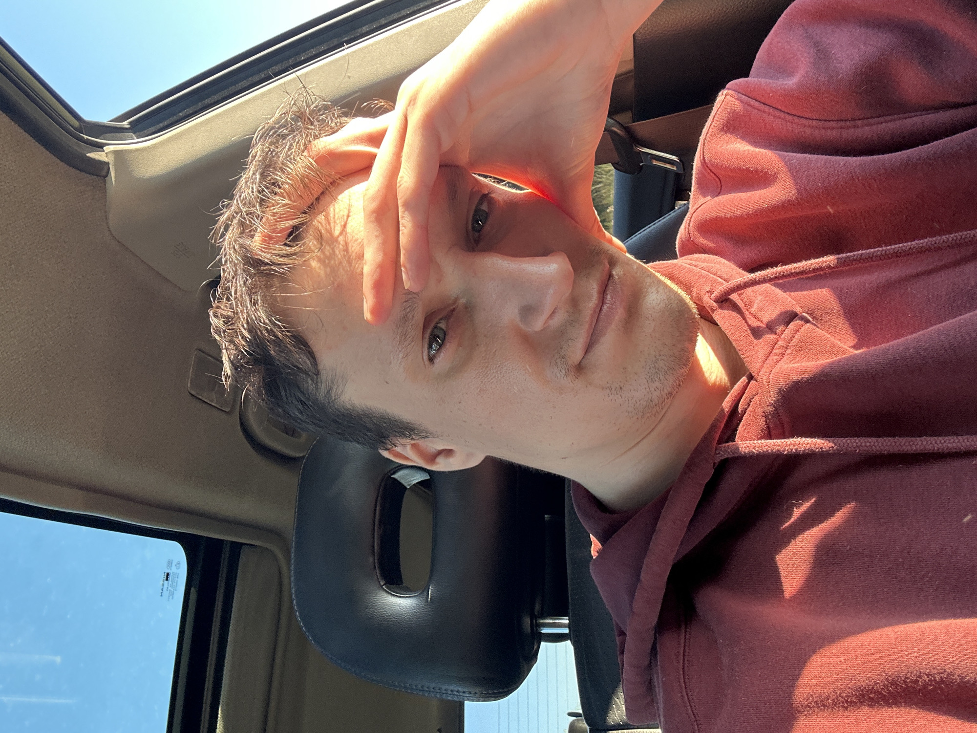 A young adult male with light brown hair is taking a selfie inside a car. He is wearing a maroon hooded sweatshirt. His right hand is placed on his forehead, and he is looking at the camera with a neutral expression. The car has a sunroof, which is open, allowing sunlight to illuminate the scene. The interior of the car, including the black headrest and a beige ceiling, is visible. The background shows a clear blue sky. The location is not identifiable as Makiv, Ukraine based on the visual information provided.