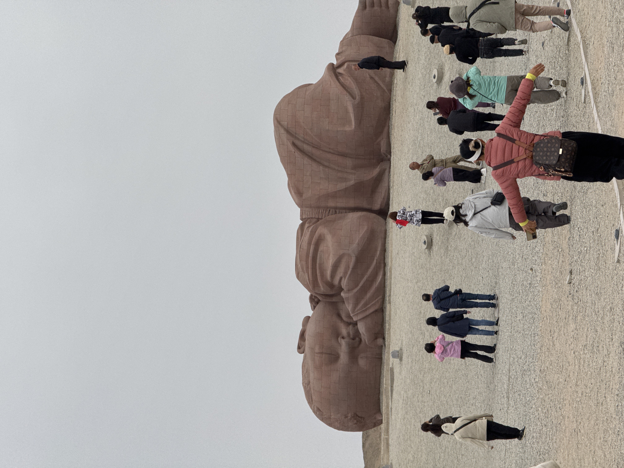 The image shows a large, horizontally positioned stone statue of a reclining baby in Guazhou County, China. A group of people, presumably tourists, stand beside it. Some individuals are looking at the statue, while others are taking pictures. The sky appears overcast. The ground is a gravel-like surface.