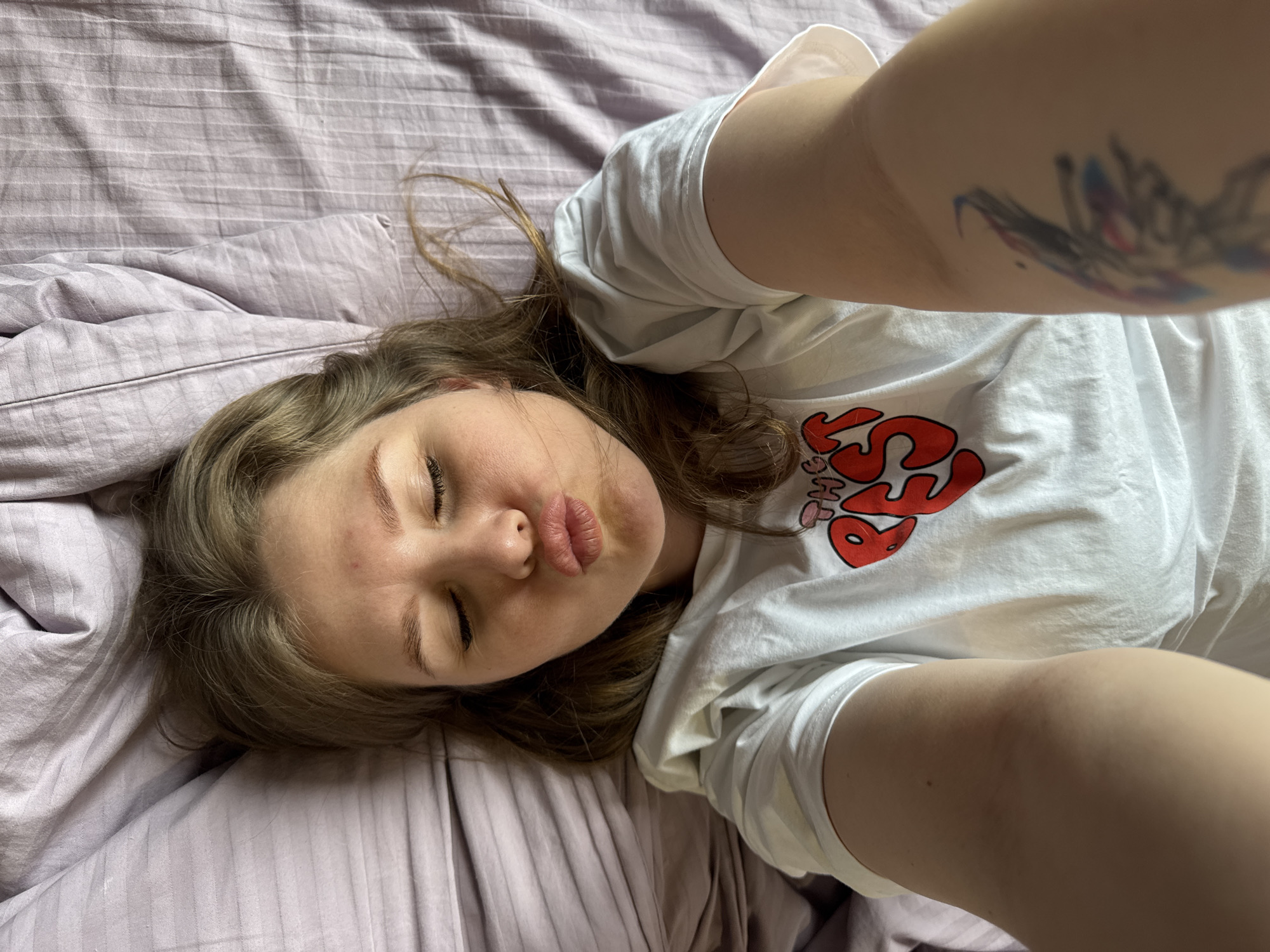 A young woman is depicted lying on a striped, light purple bedspread, taking a selfie from an overhead perspective. She has long, light brown hair fanned out around her head. Her eyes are closed, and her lips are slightly pursed in a 'kiss' or 'duck face' expression. A small red blemish is visible on her left cheek. She is wearing a white t-shirt with a prominent red and white graphic design on the chest that clearly reads "THE KISS". Her right arm is extended, holding the camera, and a colorful, abstract tattoo with blue and red elements is visible on her right upper arm/shoulder area. Part of her left arm is also visible. The lighting appears to be natural and bright, suggesting it is daytime. The setting is indoors, likely a bedroom. The overall mood is relaxed and playful. While the provided context indicates the location is Gornyi (Dvurechenskoe S/P), Russia, there are no specific visual cues within the image to confirm this geographical location.