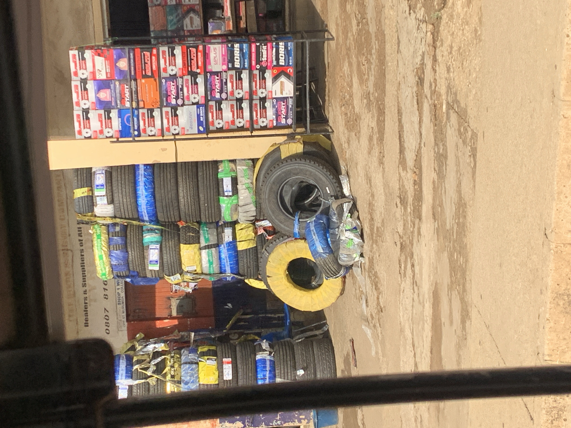 The image depicts an outdoor retail space likely located in New Stadium, Nigeria, as indicated by the "OPP ST. MURUMBA COLLEC" on a sign that partially reads "Denters & Suppliers of All" and a phone number starting with "0807 816." The main focus is on a display of tires stacked vertically and banded with colorful wrapping, presumably for sale. Above the tires is a shelf holding numerous car batteries in boxes, stacked and arranged. Brands such as "SUPER START", "BORIS," "SOLITE," and "UPLUS" are visible on the battery boxes. The surrounding environment appears to be a commercial area with a rough, concrete surface nearby. The image is taken from a slightly elevated position. No people are directly visible in the image.