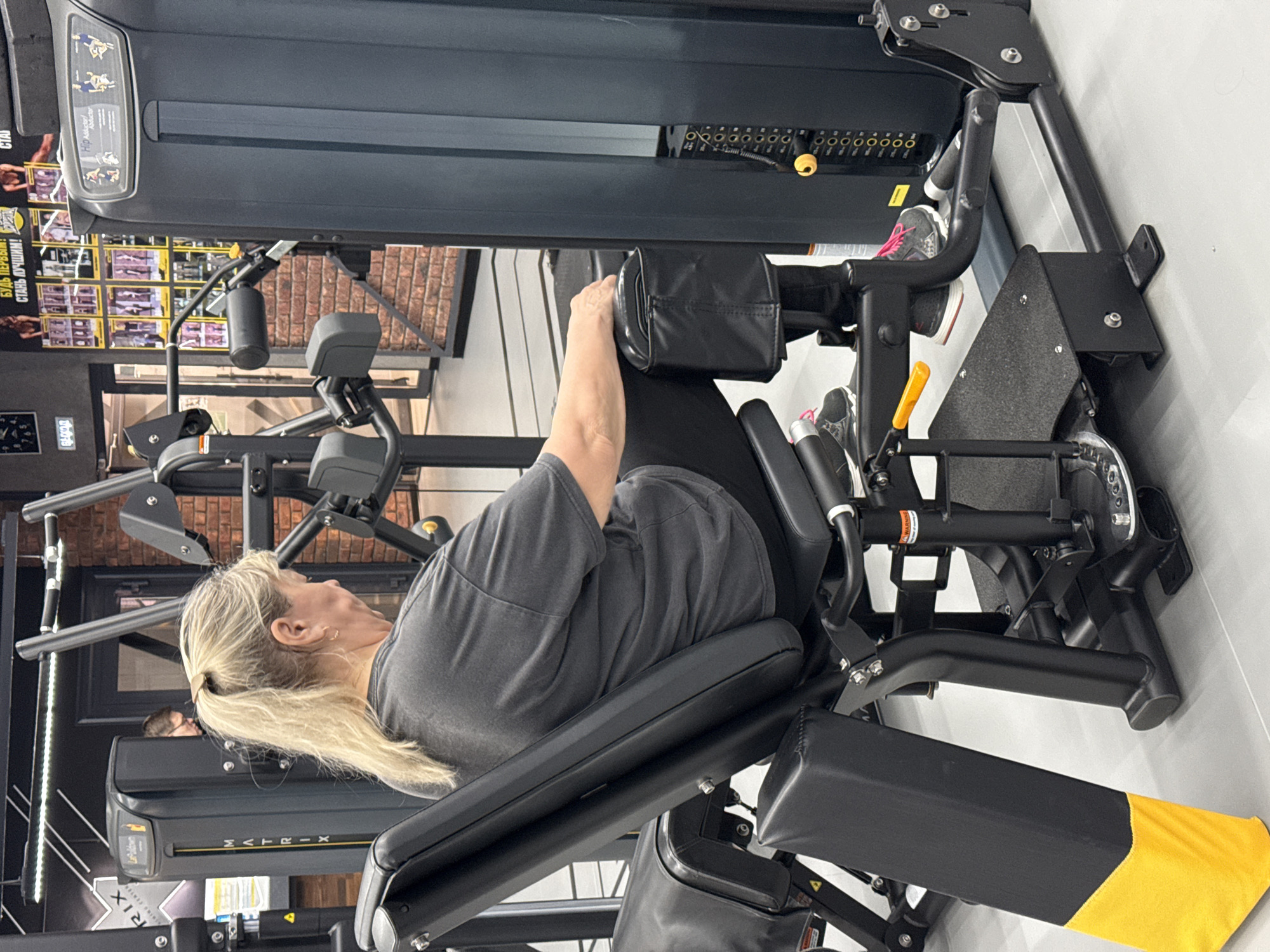 A woman with blonde hair pulled back in a ponytail is positioned on a seated leg press machine in a gym. She is wearing a grey t-shirt. The machine is primarily black with some silver components, and has black padded supports for her back and legs. A weight stack is visible at the top of the machine. The gym has exposed brick walls, grey flooring, and other exercise equipment in the background. The location is Votkinsk, Russia.
