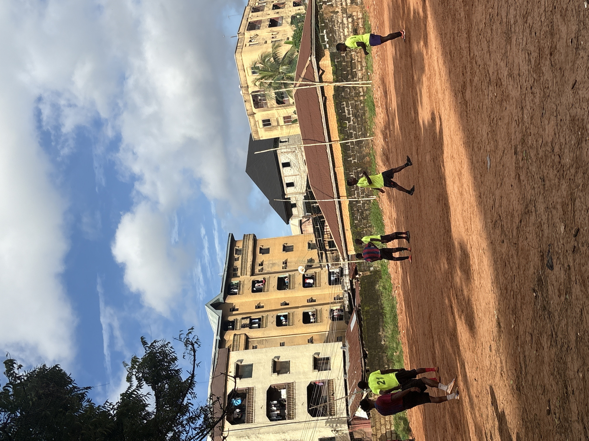 This is a street-level, medium-angle, outdoor shot taken on a dusty, red-dirt soccer field in Onitsha, Nigeria. The sky is a vibrant blue, dotted with fluffy white clouds. The background features a row of apartment buildings, predominantly yellow and beige, with balconies adorned with laundry. Several young men, dressed in soccer attire, are engaged in a game. One player in a yellow jersey with the number "21" is visible, alongside others in similar bright jerseys and dark shorts, and one in a striped jersey. The field itself is uneven and dry, with scattered debris. The scene suggests a casual, local sporting activity taking place amidst the urban landscape of Onitsha.